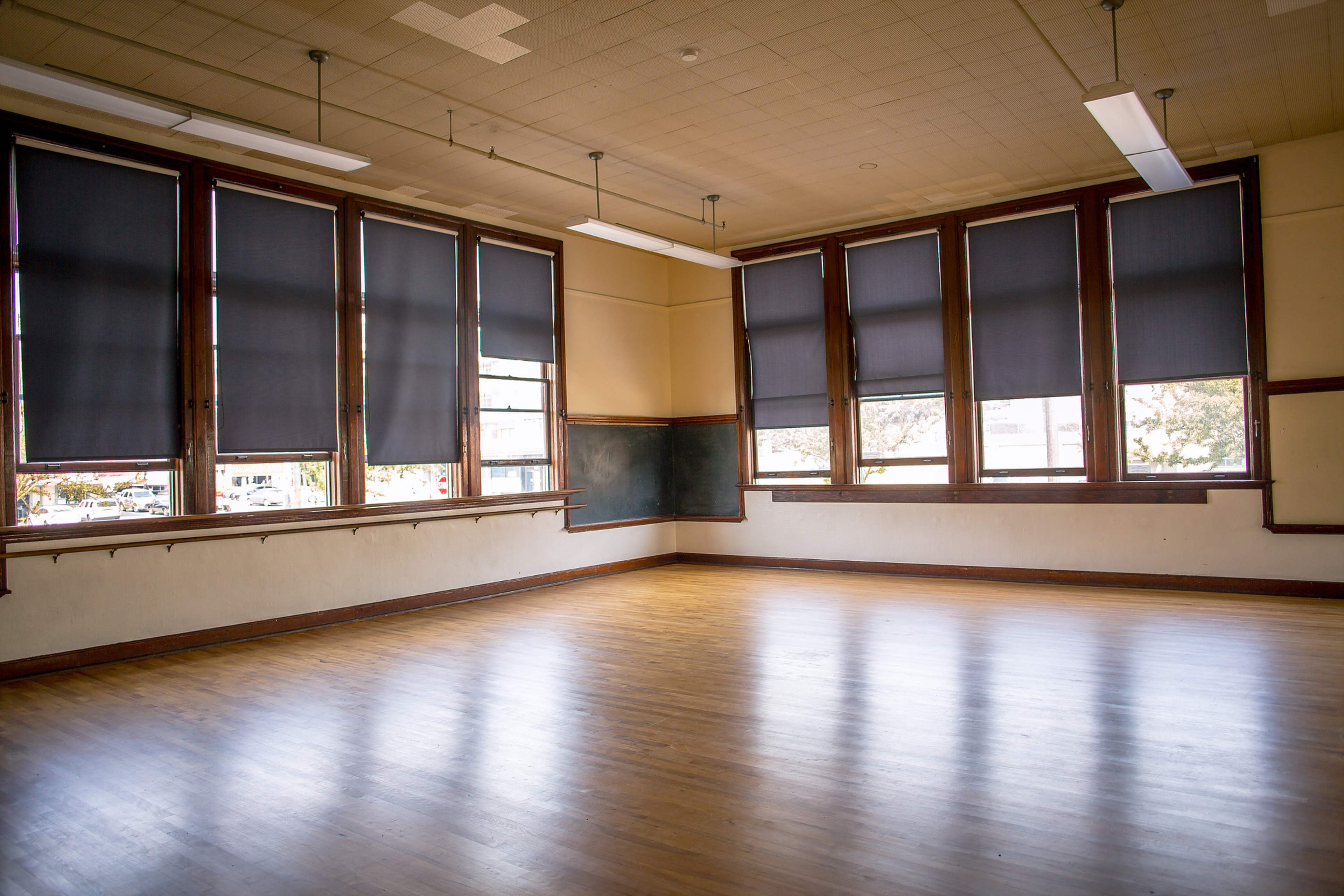 An empty classroom with large windows covered by dark shades and a wooden floor.