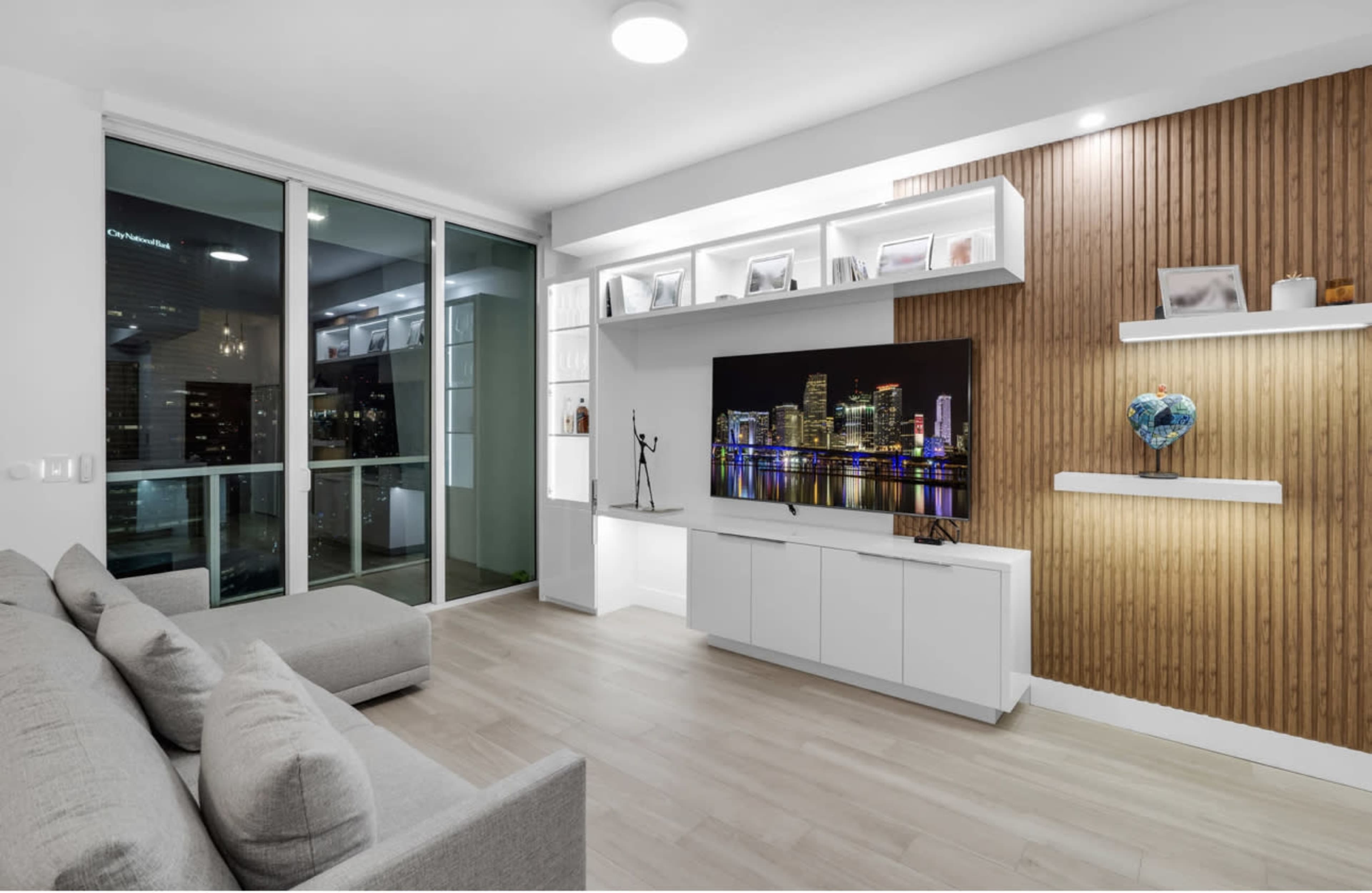 The scene shows a modern living room with a large television mounted on a wooden accent wall, white shelves displaying decor, and a gray sofa facing the TV.
