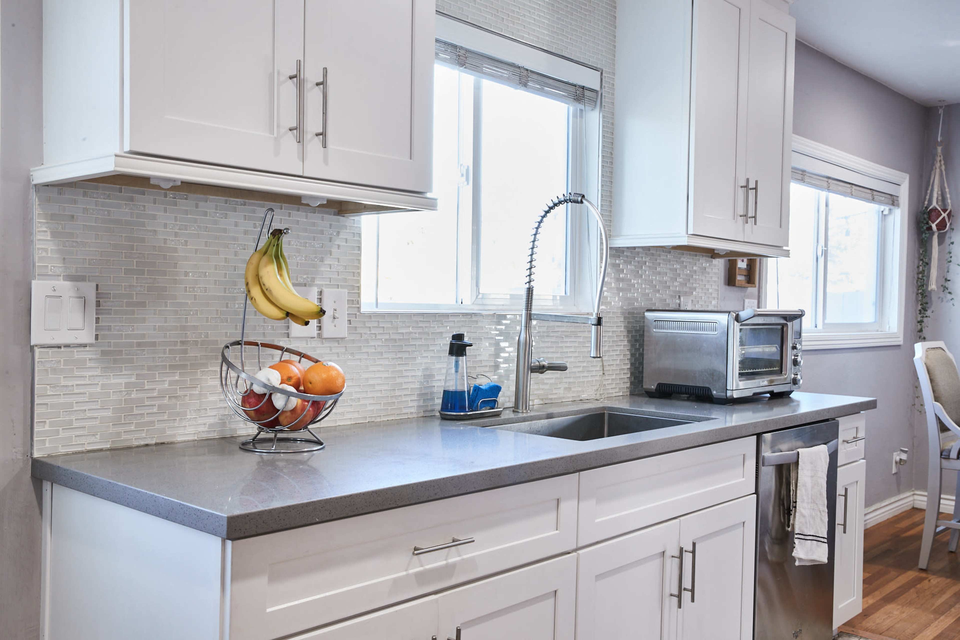 A modern kitchen features white cabinetry, a gray countertop, and a stainless steel sink with a window above, along with a fruit basket and a toaster visible on the counter.