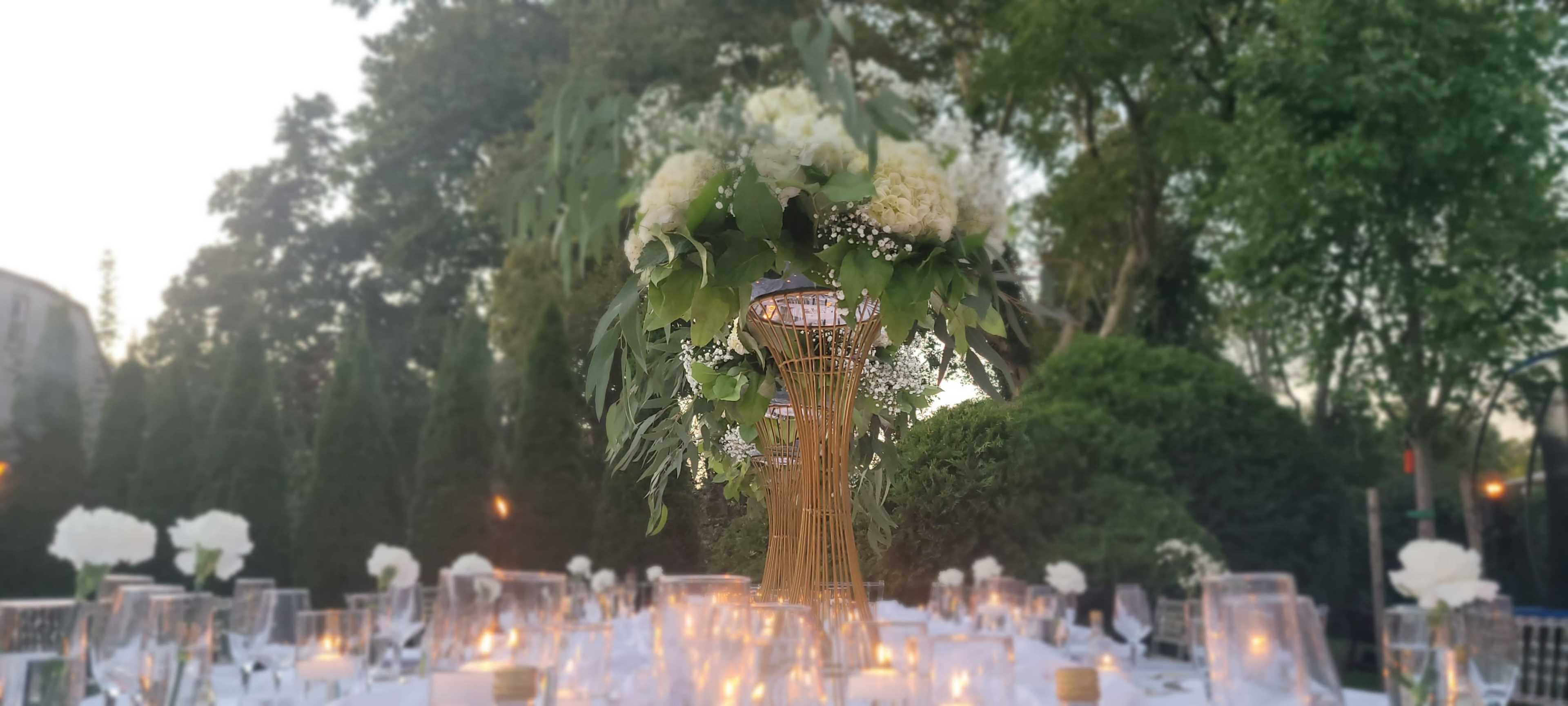 A beautifully arranged table centerpiece features a tall floral display made of white flowers and greenery at an outdoor event.