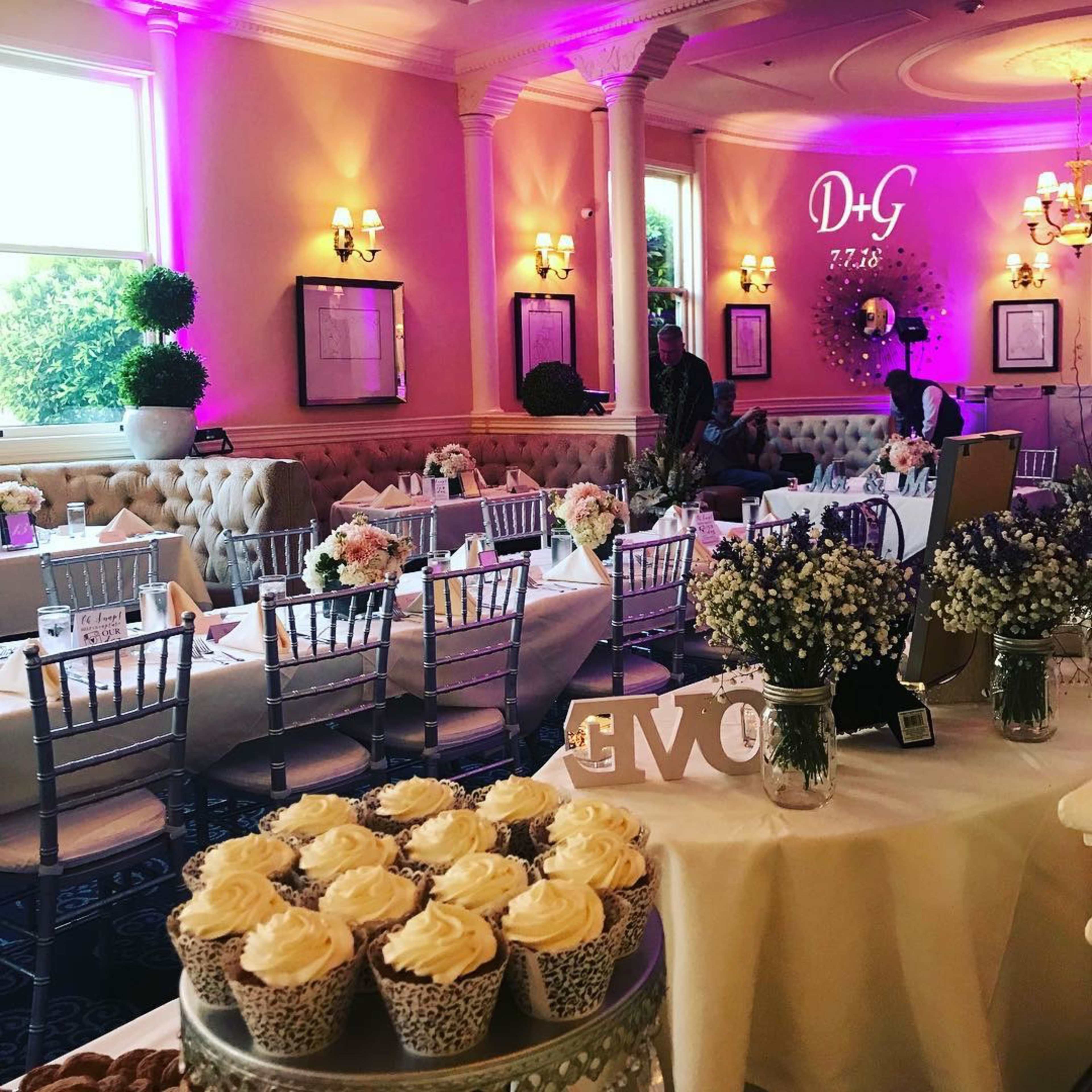 A decorated event space with elegantly set tables, floral centerpieces, and a display of cupcakes, illuminated by soft purple lighting.