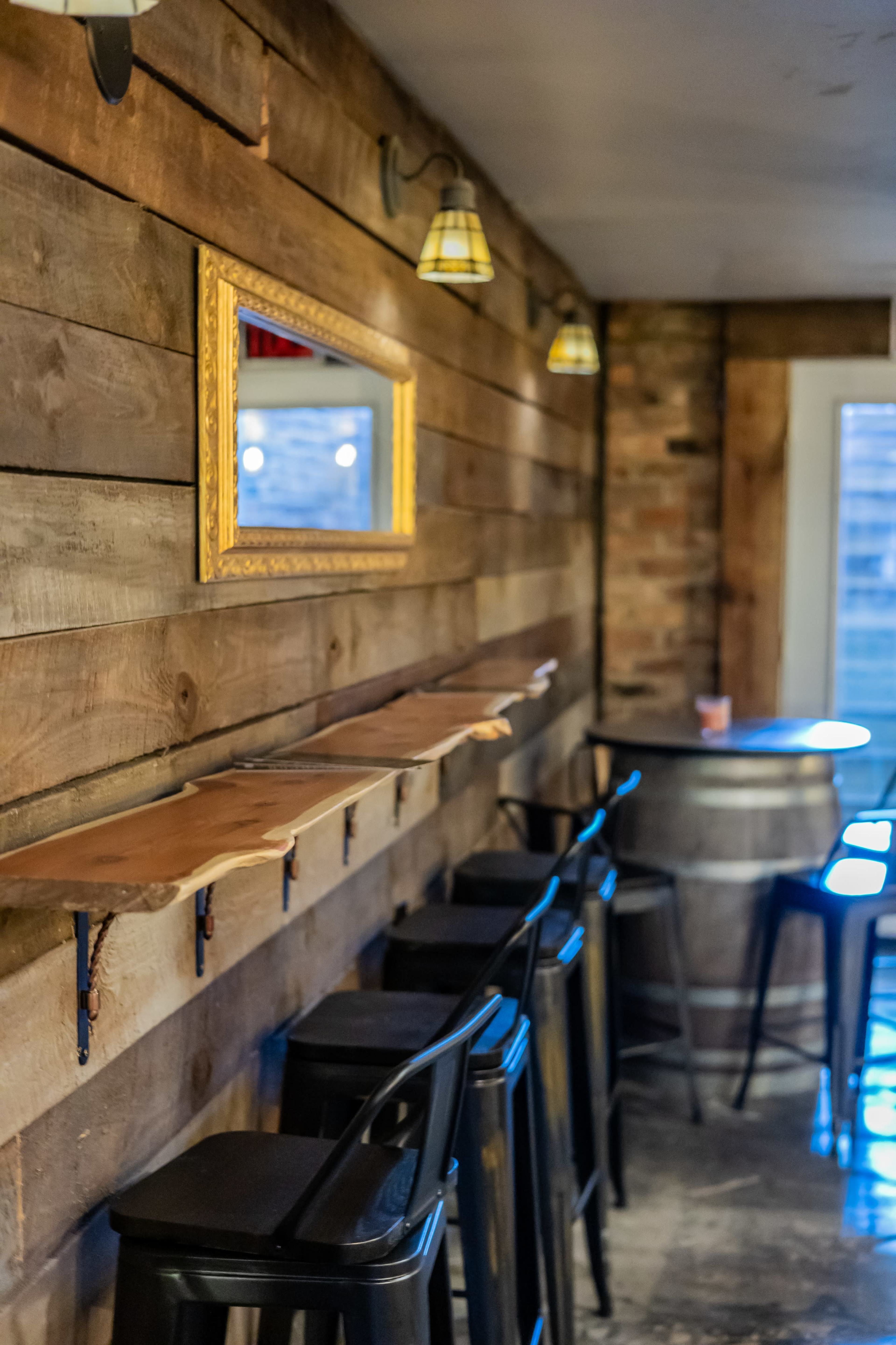 The image features a rustic interior with wooden walls, a mirror framed in gold, a long wooden shelf, black bar stools, and a barrel table.