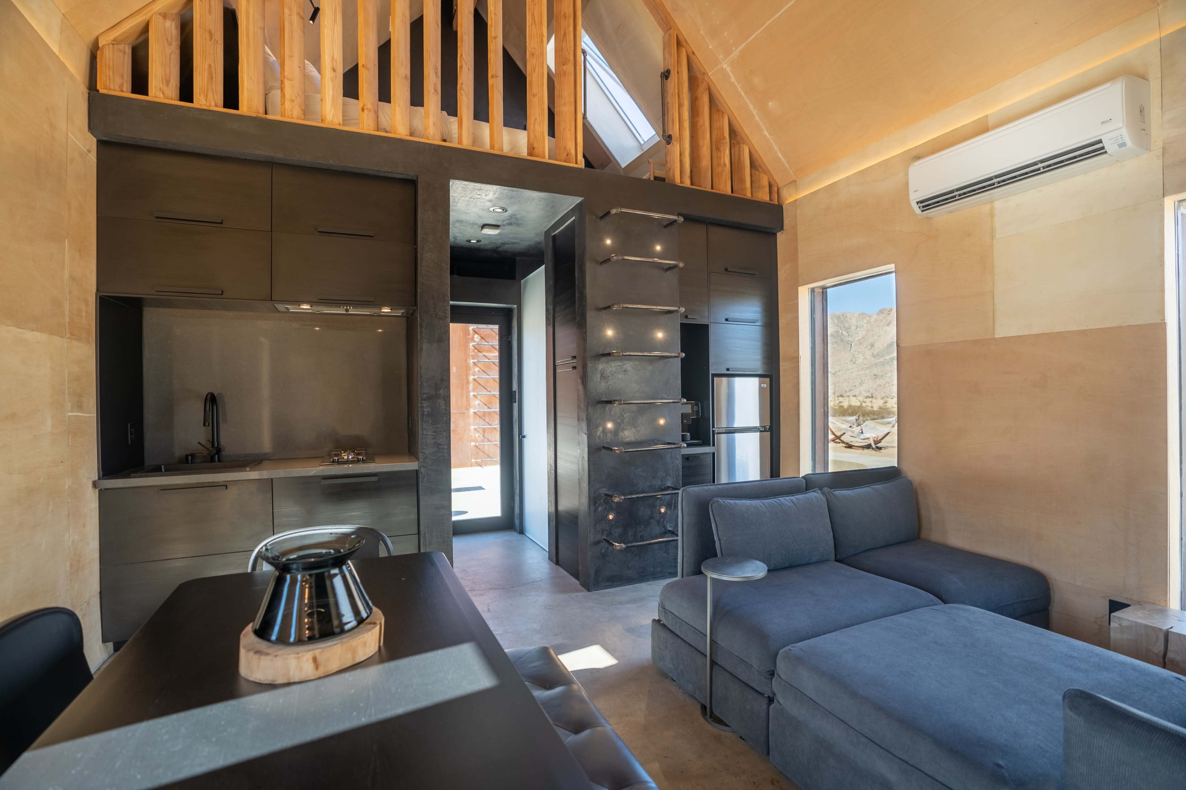 The interior of a modern, compact cabin features a kitchen area with dark cabinetry, an open living space with a sectional couch, and large windows showcasing a mountainous landscape.