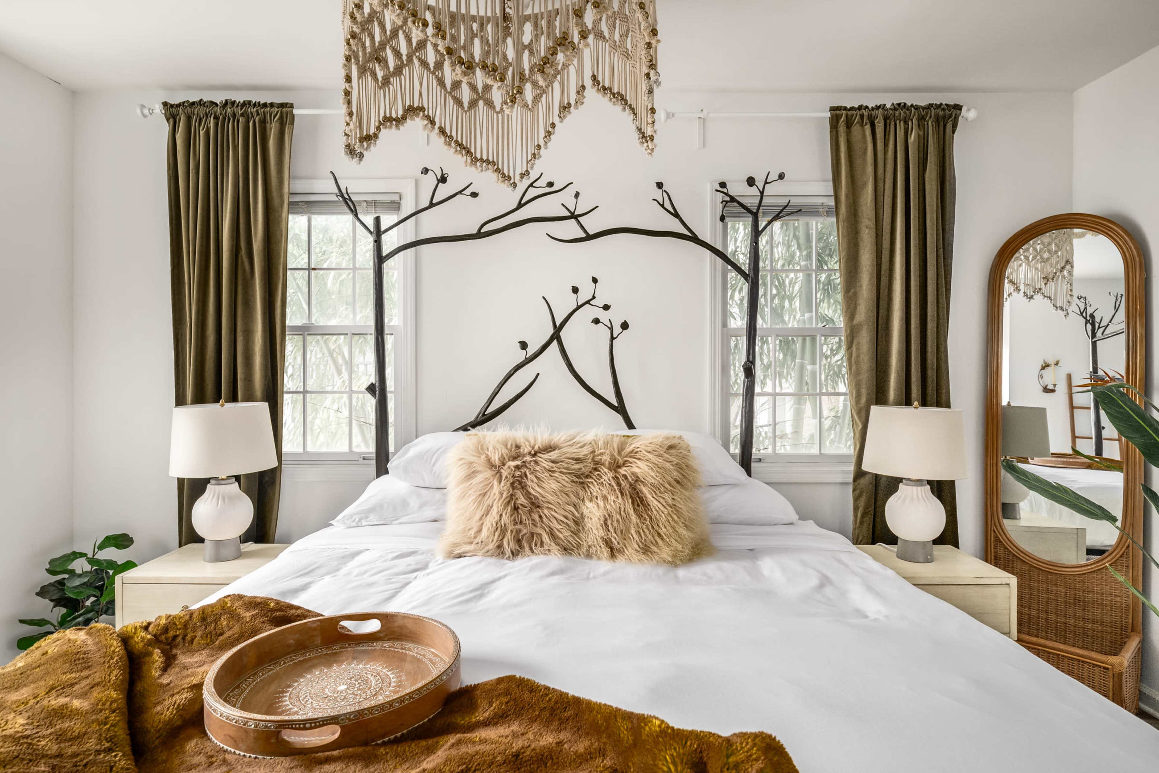 The image shows a neatly arranged bedroom featuring a bed with a white comforter, two bedside lamps, and decorative wall art resembling branches, along with green curtains and a mirror.