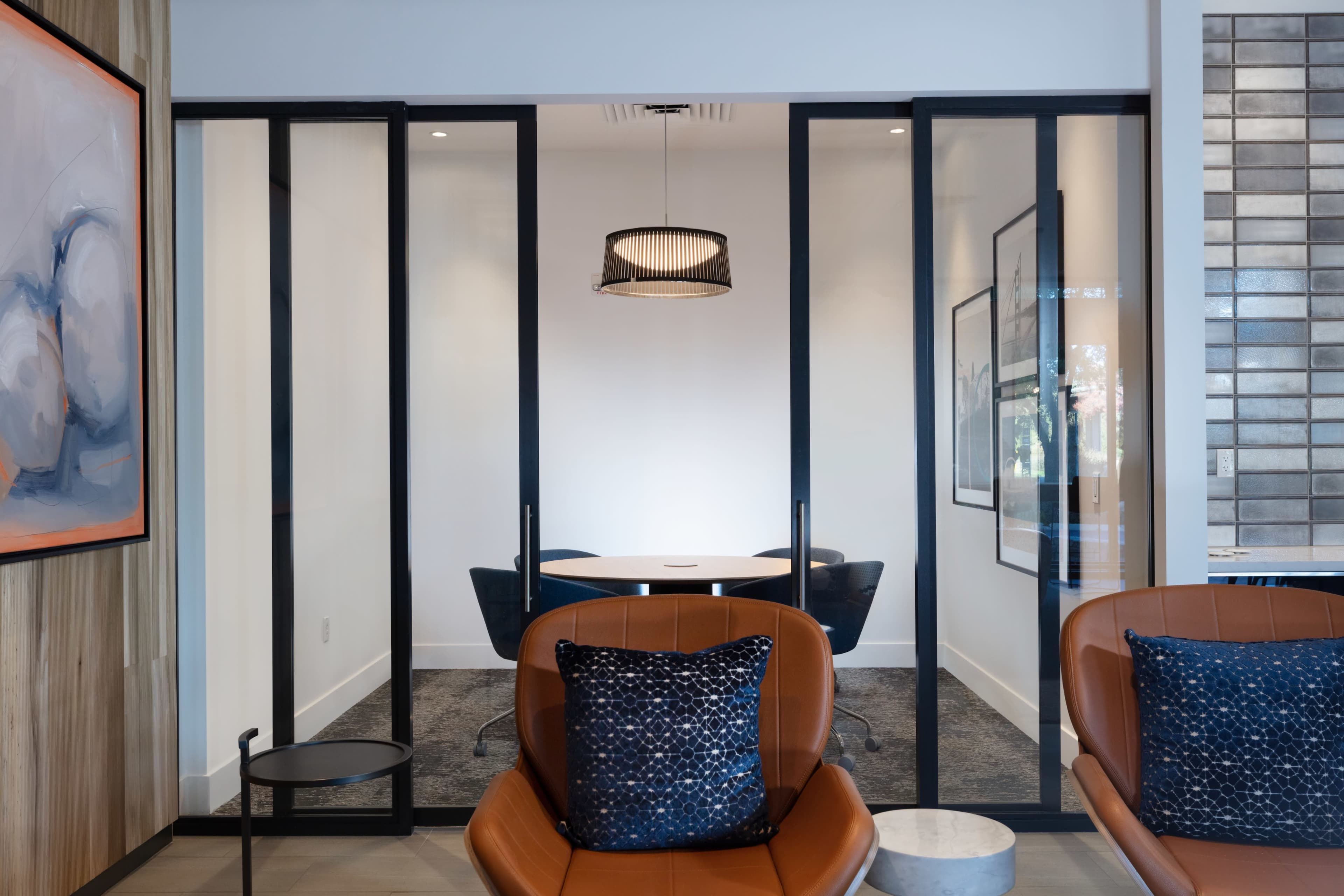 A modern office space featuring a glass-enclosed meeting room with a round table and black chairs, accompanied by orange leather seating in the foreground.