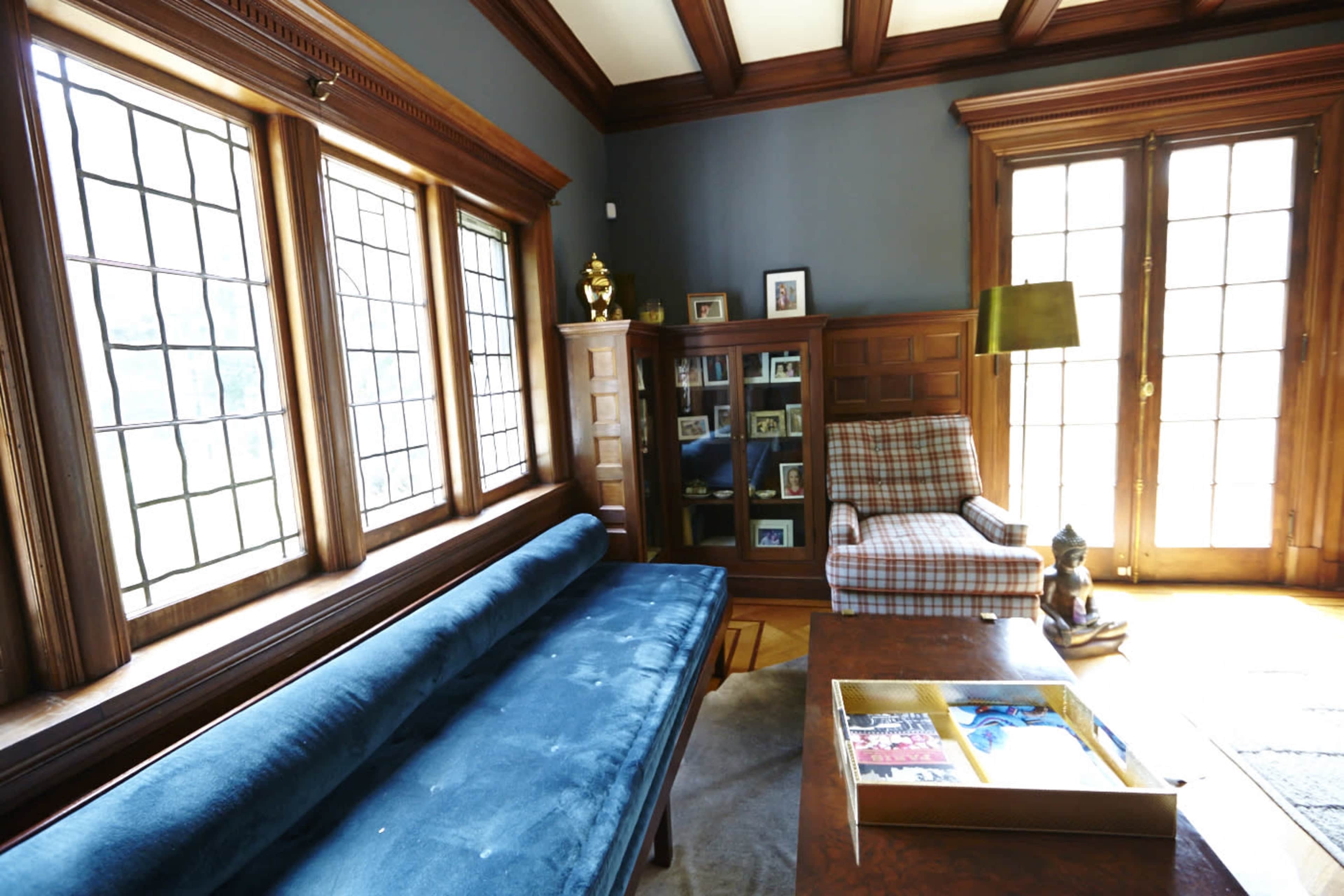The room features a blue sofa, wooden cabinetry with bookshelves, a plaid armchair, and large windows along one wall.