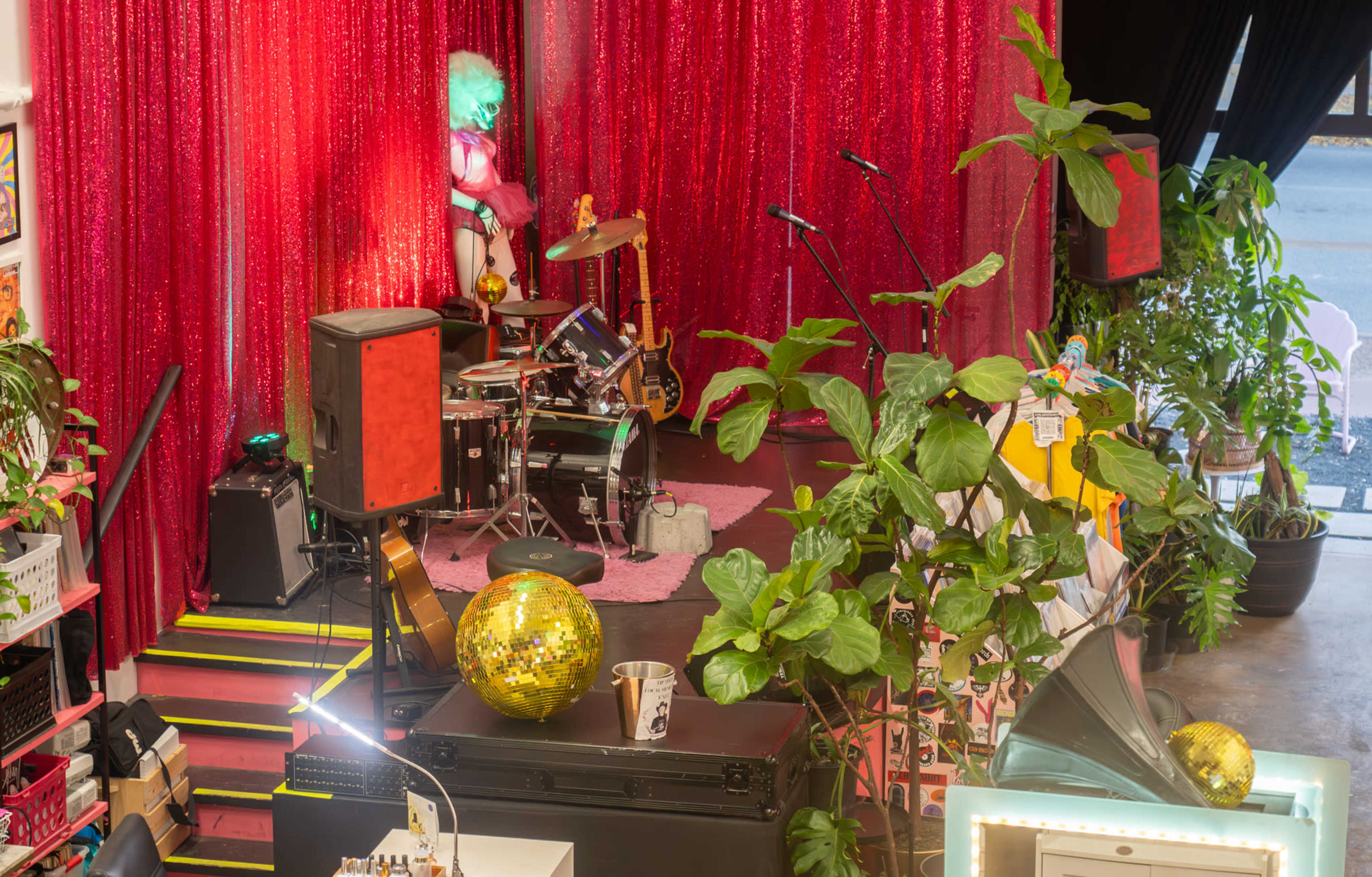The image shows a performance space with a glittery red backdrop, a small drum set on a pink rug, and various potted plants surrounding the area.
