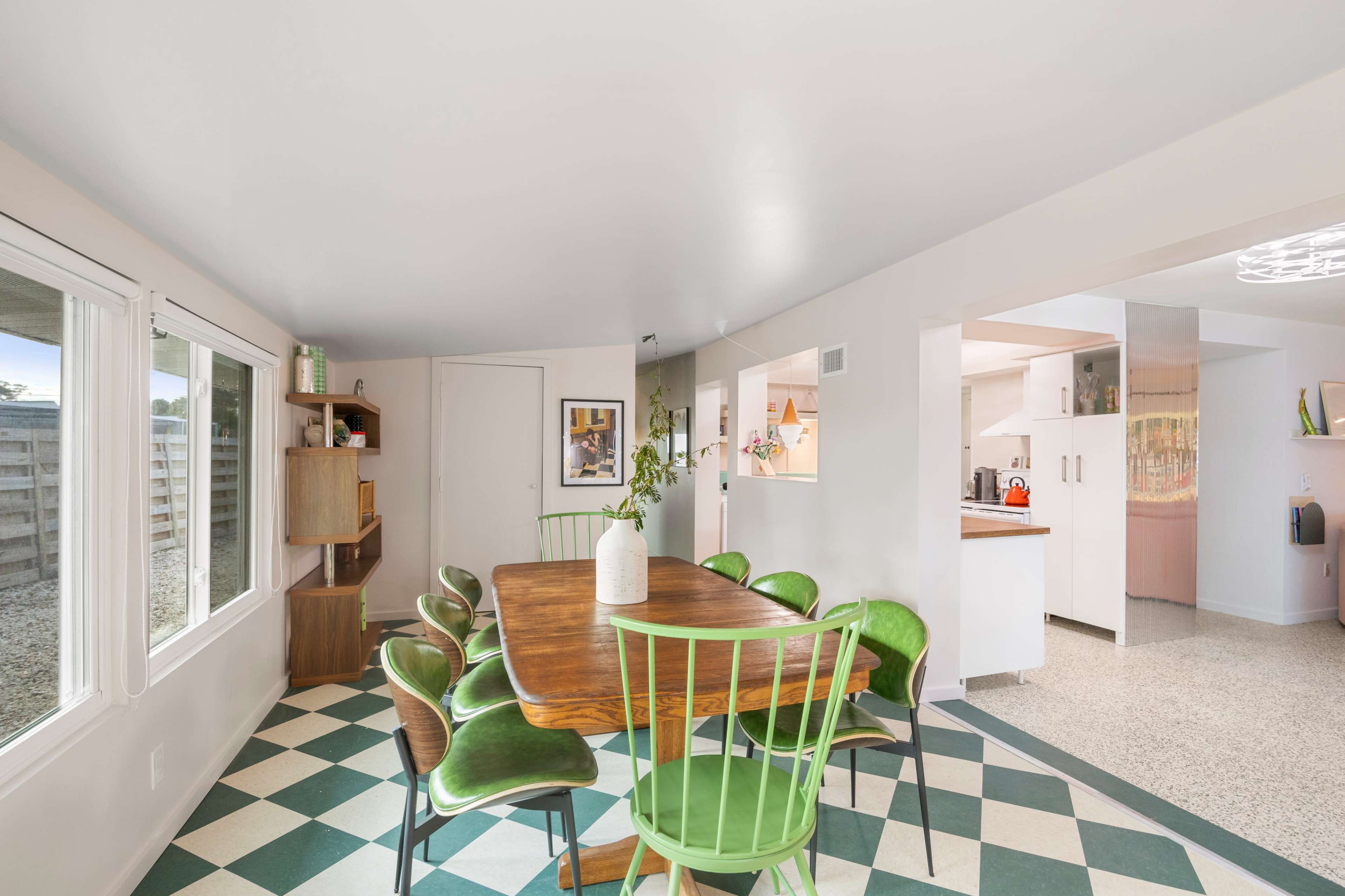 A dining area features a wooden table surrounded by green chairs, with large windows providing natural light and a glimpse into an adjoining kitchen.