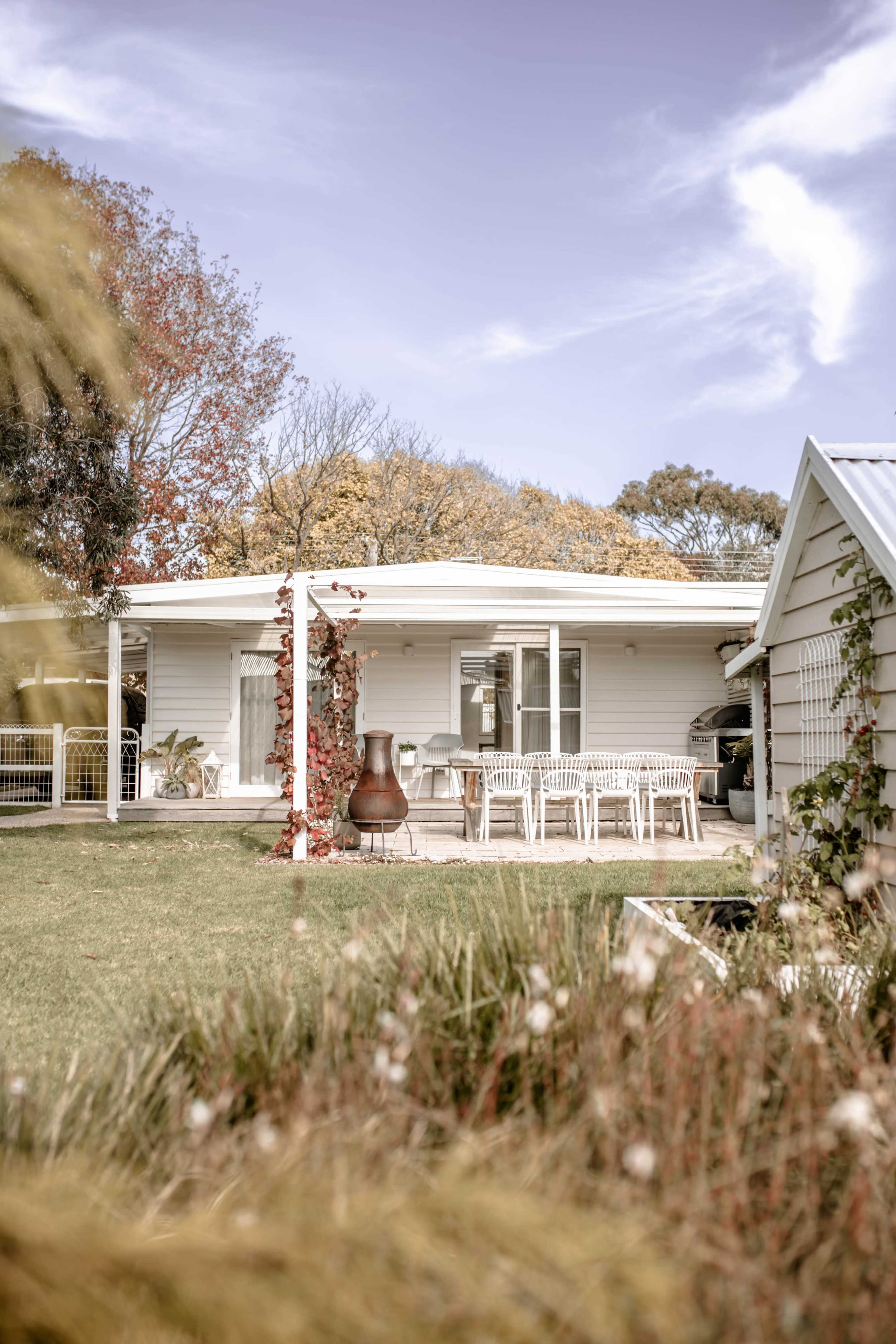 A light-colored house with large windows and a patio containing a dining table is surrounded by greenery and autumn foliage.