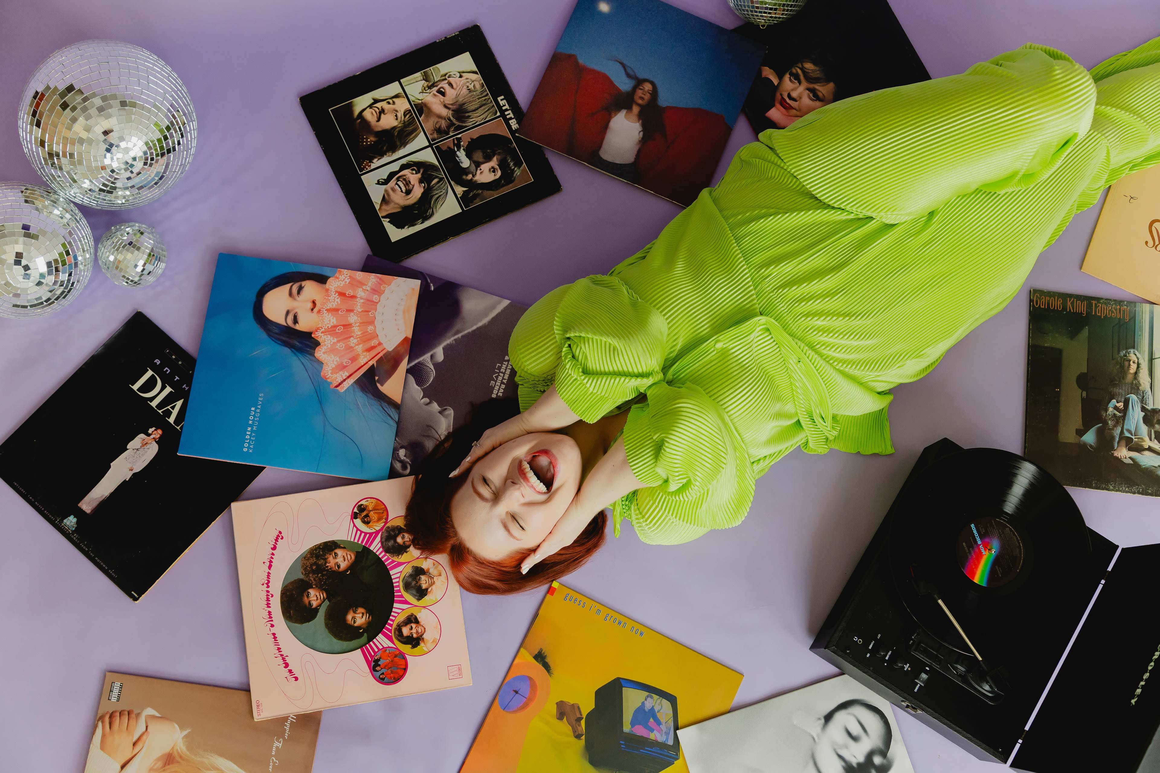 A person wearing a bright green outfit lies on a purple surface surrounded by vinyl records and a record player.