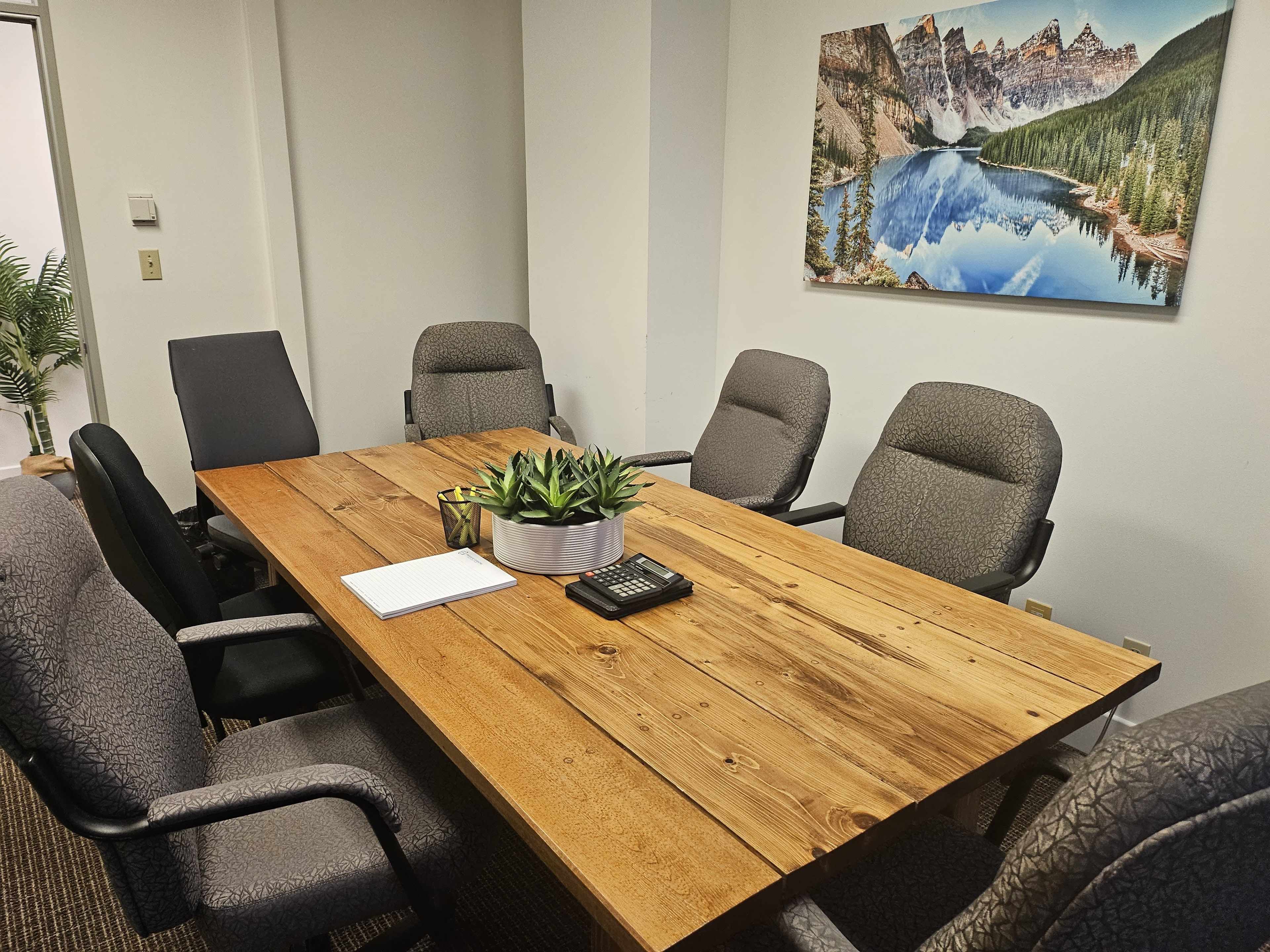A conference room featuring a wooden table surrounded by six upholstered chairs, with a plant centerpiece and a mountain landscape photograph on the wall.