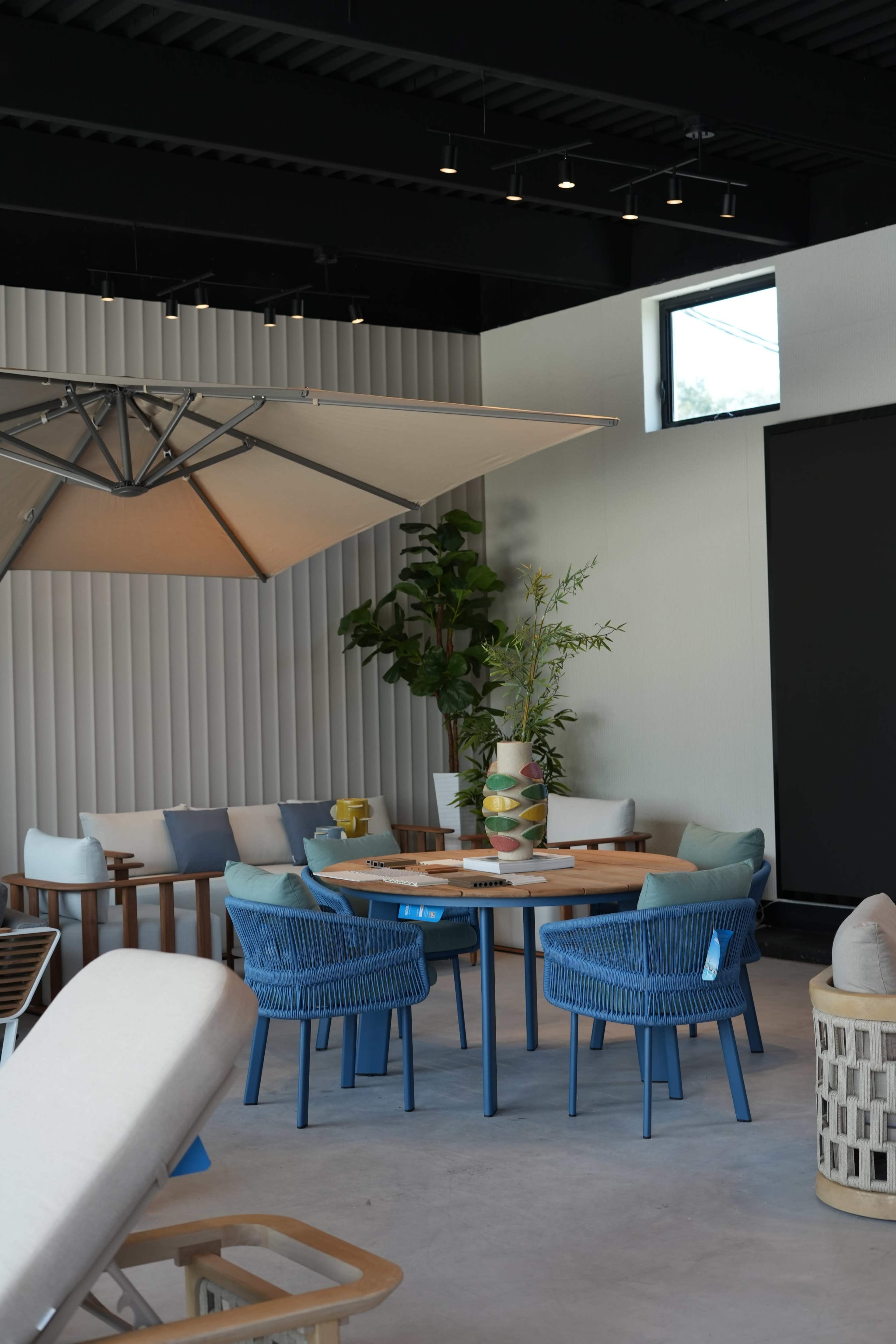 A modern indoor seating area features a round table surrounded by colorful chairs, a large umbrella, and potted plants.