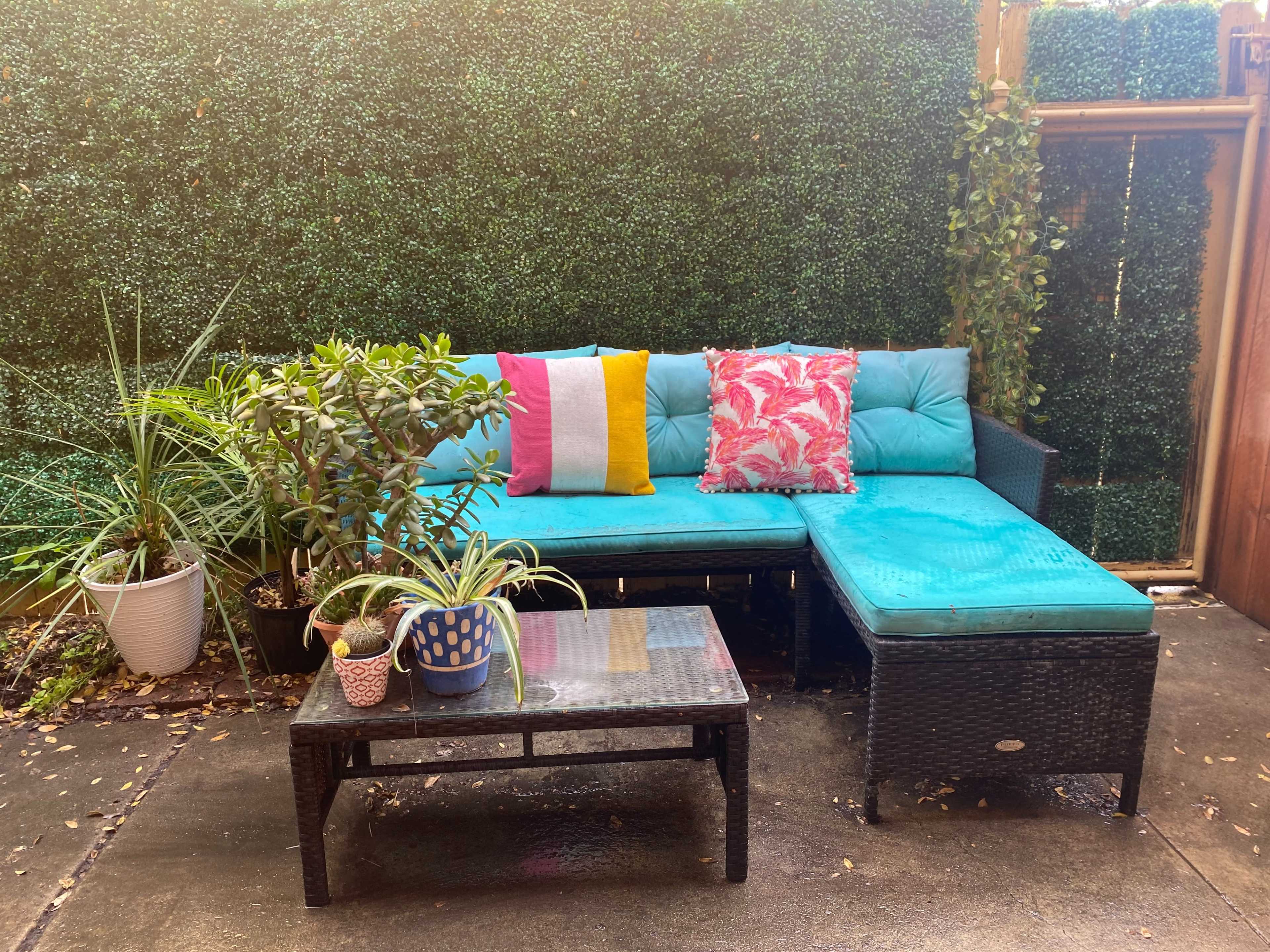 A seating area with a turquoise couch adorned with colorful cushions is surrounded by potted plants and a black coffee table on a concrete floor.