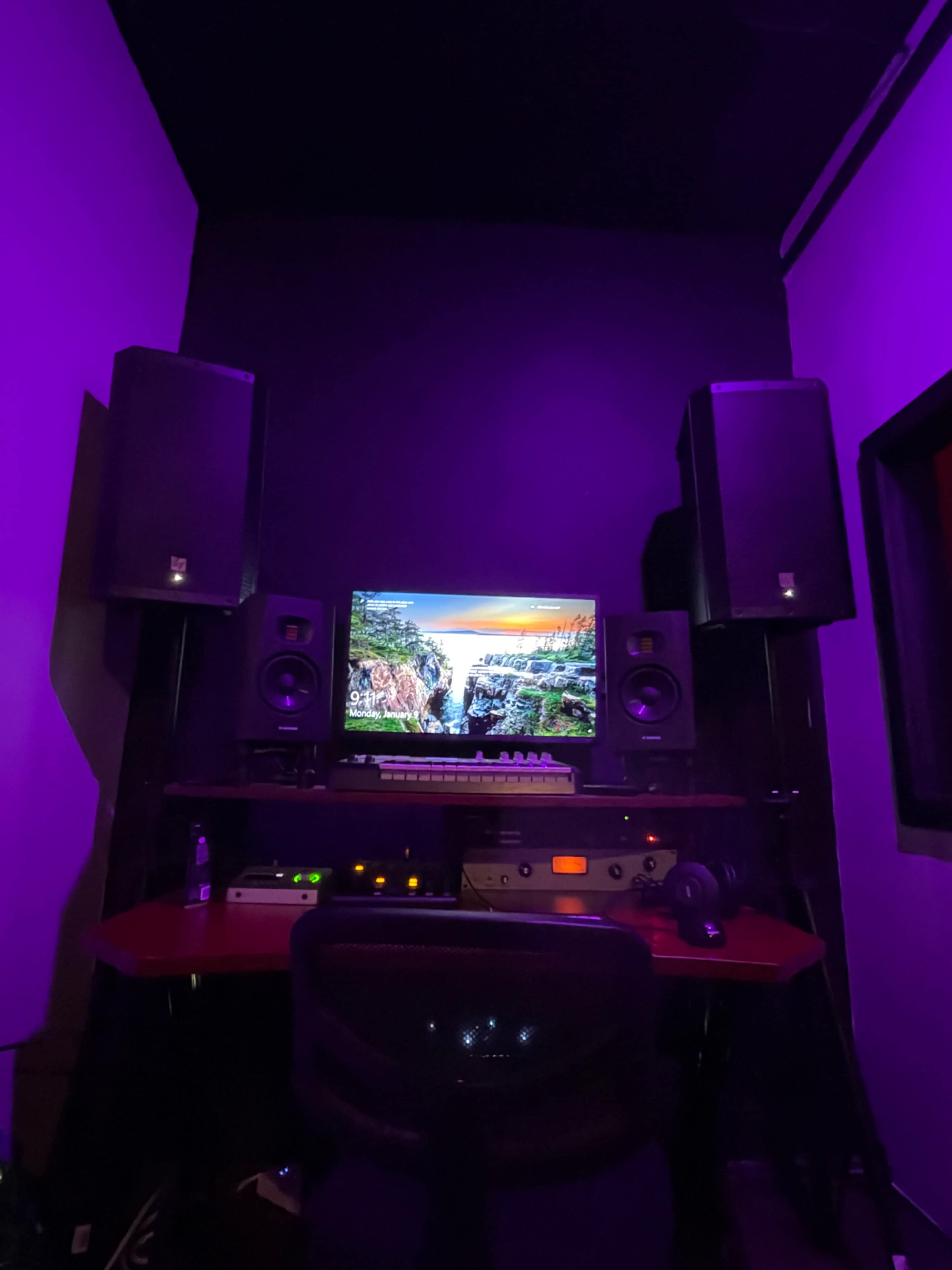 The image shows a music production studio featuring a computer monitor, audio speakers, and a red desk surrounded by dark walls.