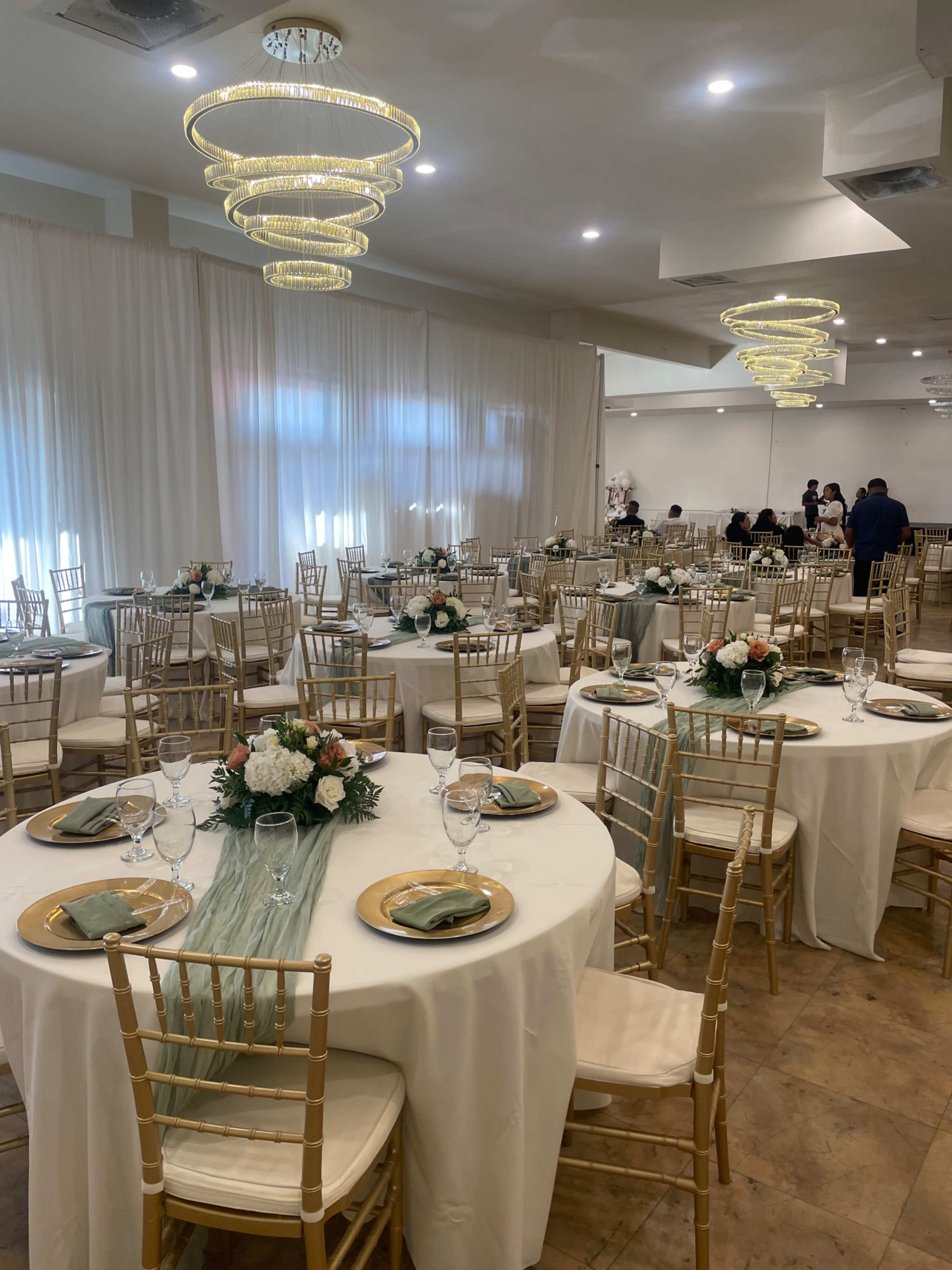 The image shows a banquet hall set up for an event, featuring round tables adorned with centerpieces, tableware, and gold chiavari chairs under decorative chandeliers.