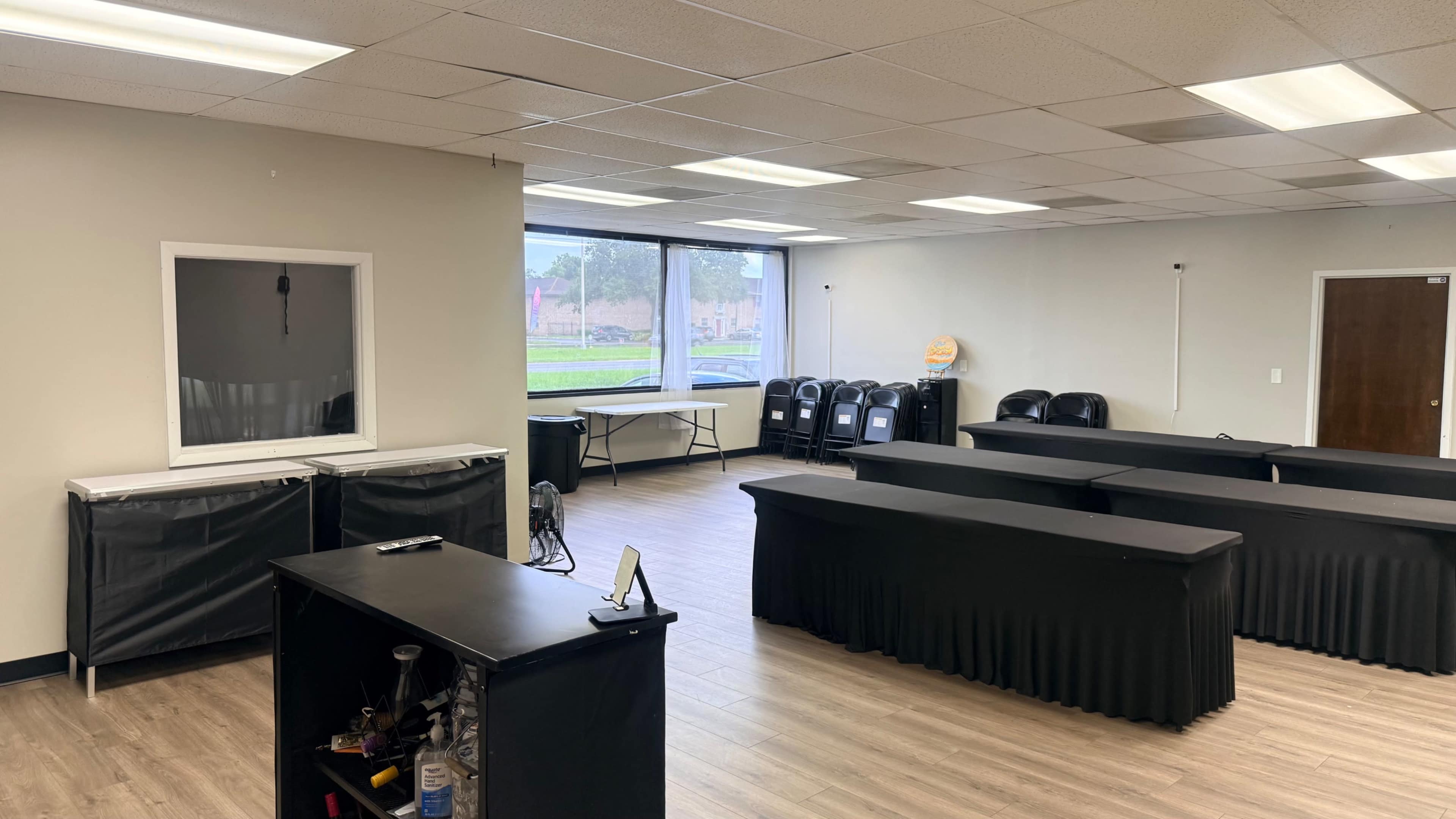The image shows a vacant room with several tables covered in black tablecloths, chairs lined up against the wall, and a large window letting in natural light.
