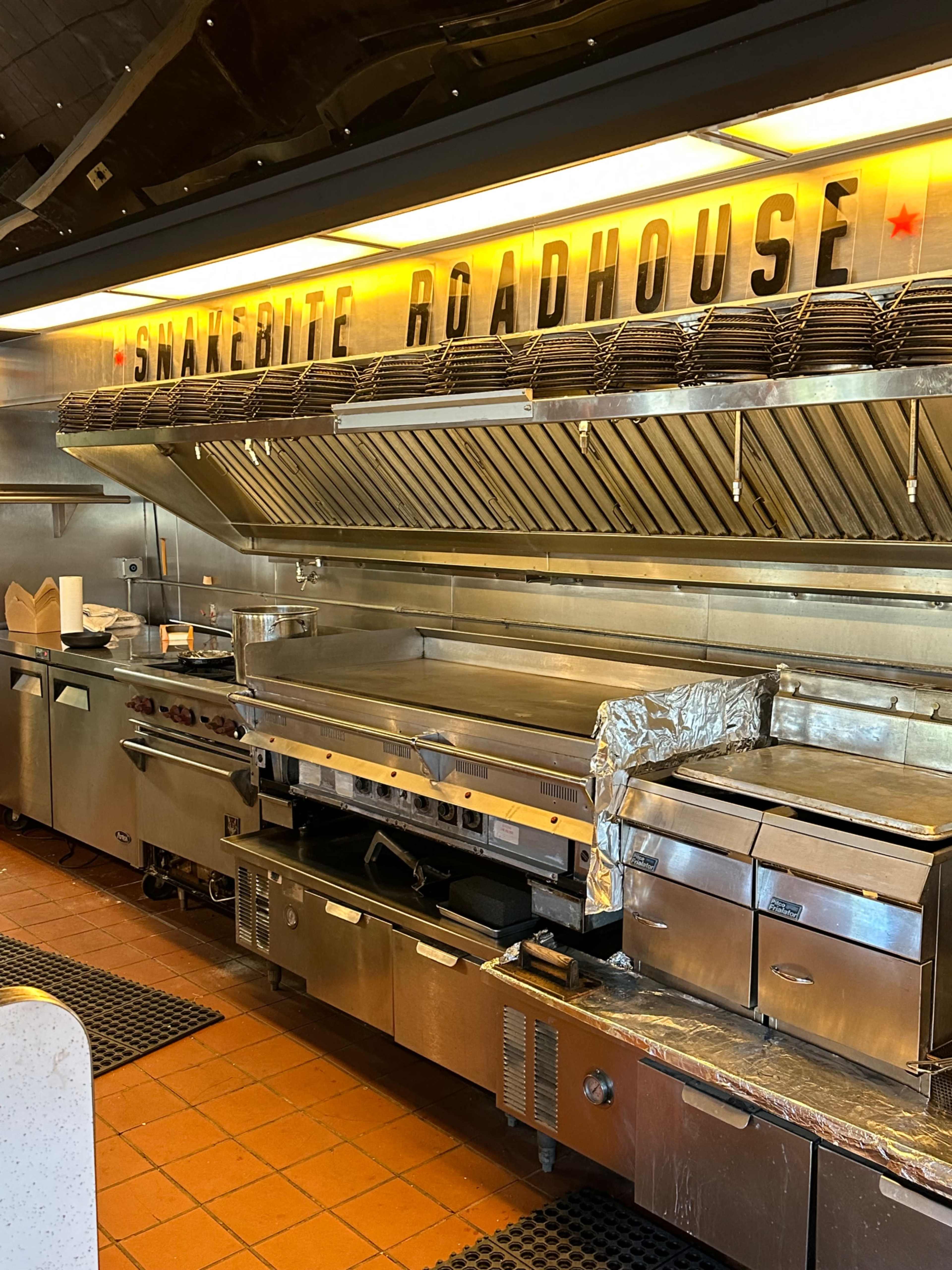 The image shows the interior of a restaurant kitchen with metallic cooking equipment and a sign reading "Snakebite Roadhouse."