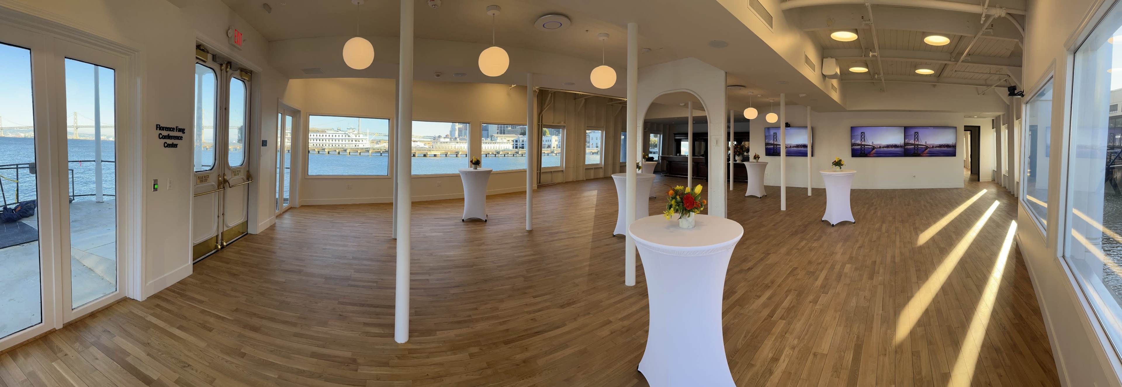 An empty event space with white tables and large windows overlooking a waterfront view.