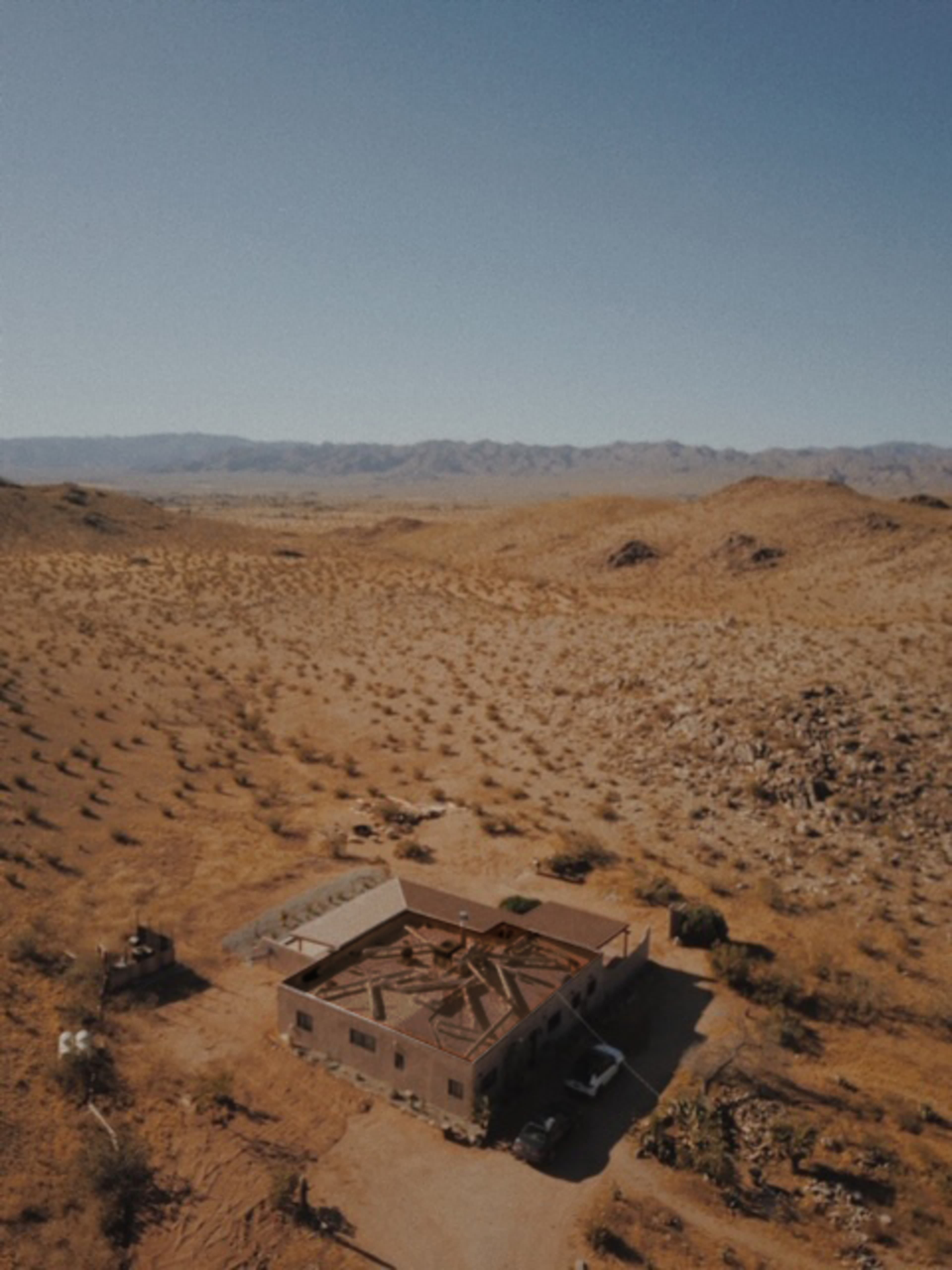 An abandoned building with a partially collapsed roof is situated in a desert landscape, surrounded by sparse vegetation and distant mountains.