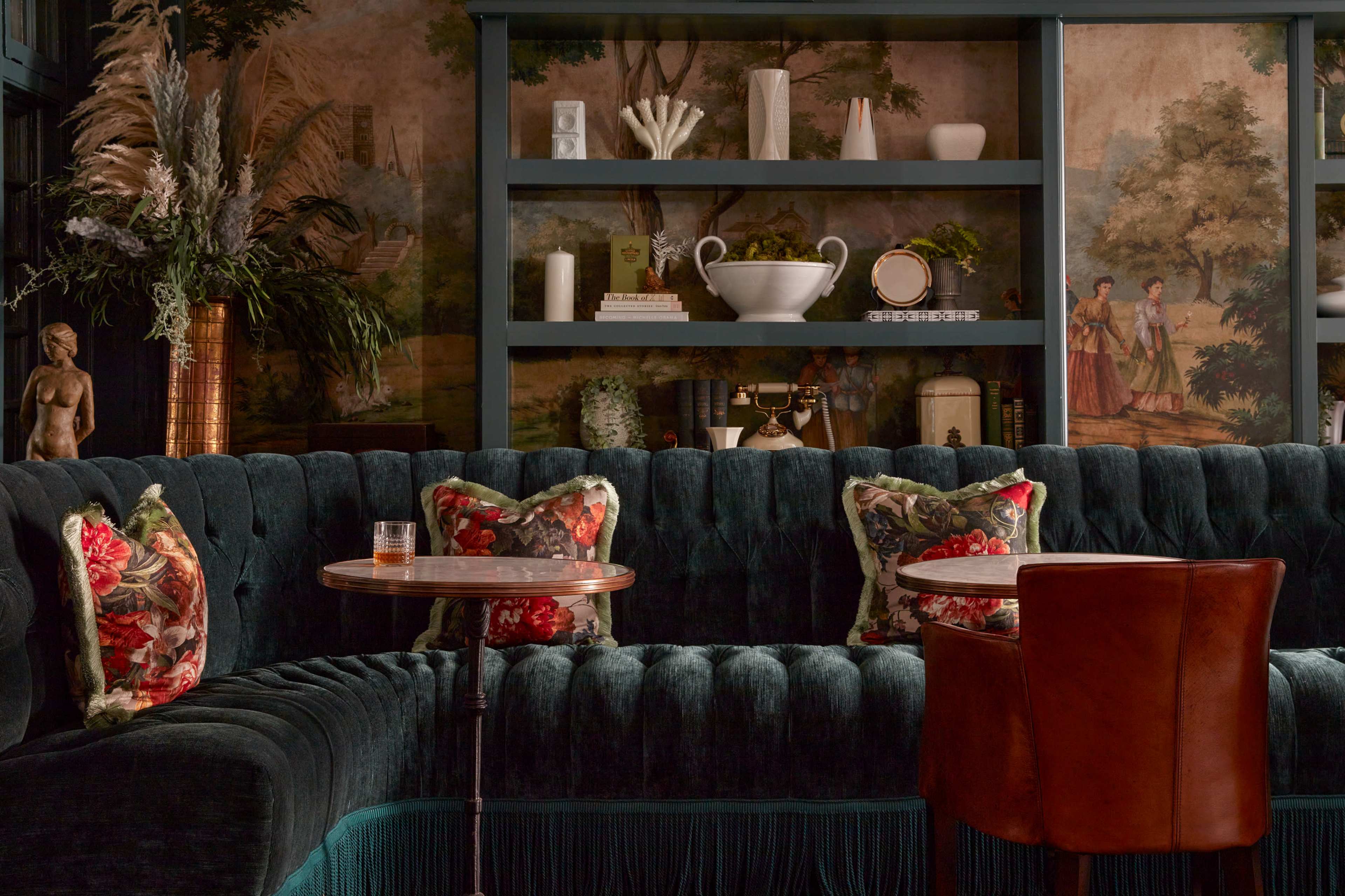 The image depicts a stylish lounge area featuring a plush green velvet sofa adorned with floral-patterned pillows, small round tables, and decorative shelves with plants and art.