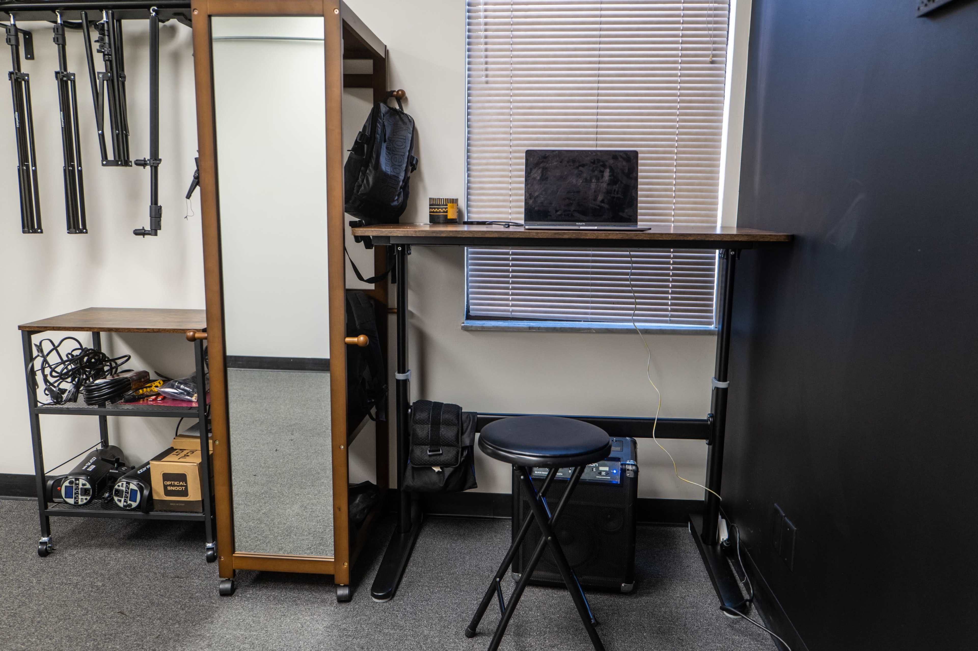 A standing desk beside a mirror, a laptop on the desk, and a stool in front of it, with storage units and equipment visible in the background.