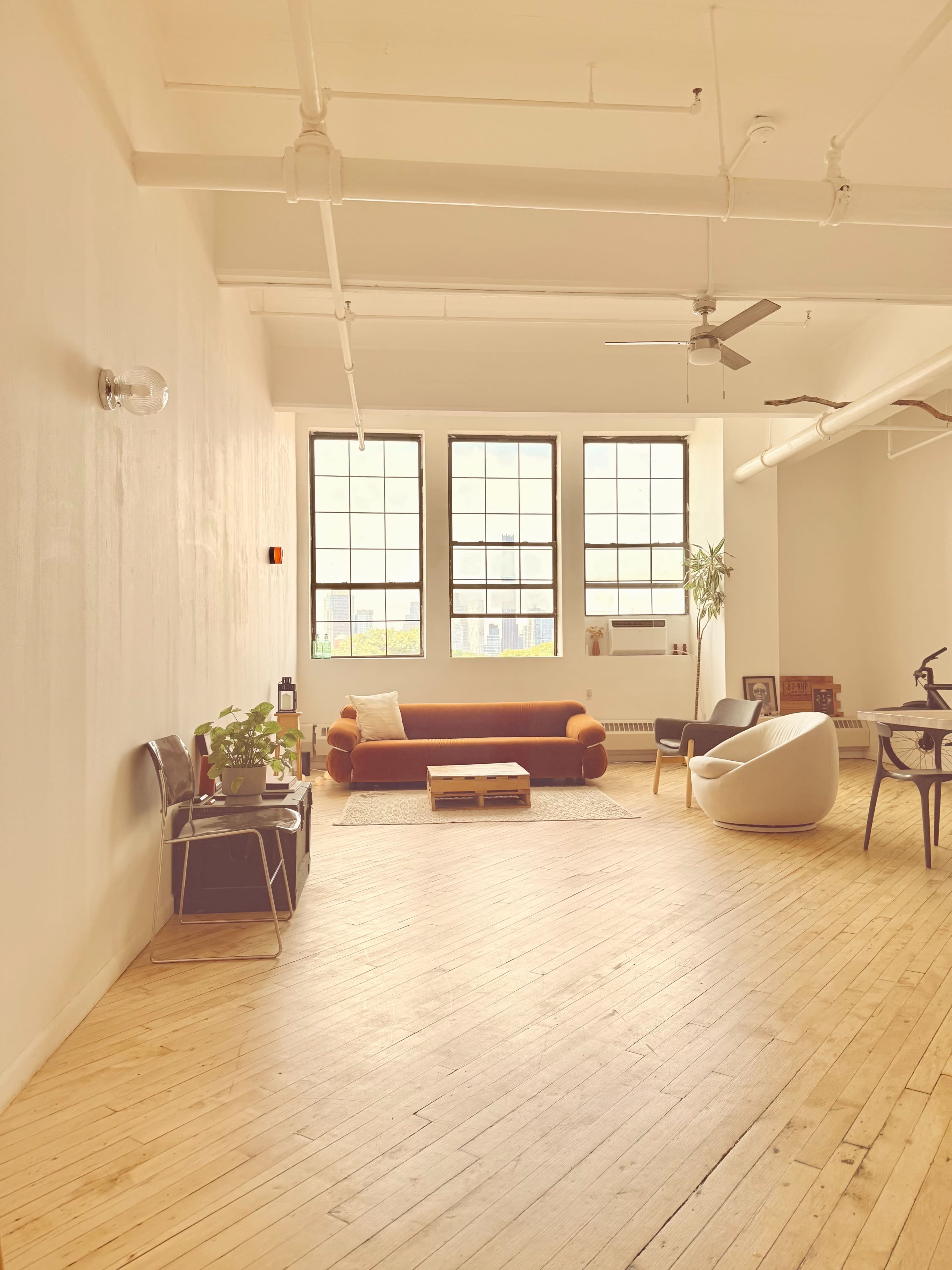 The image shows a spacious, well-lit room with large windows, featuring a brown sofa, a coffee table, a white chair, and some greenery.