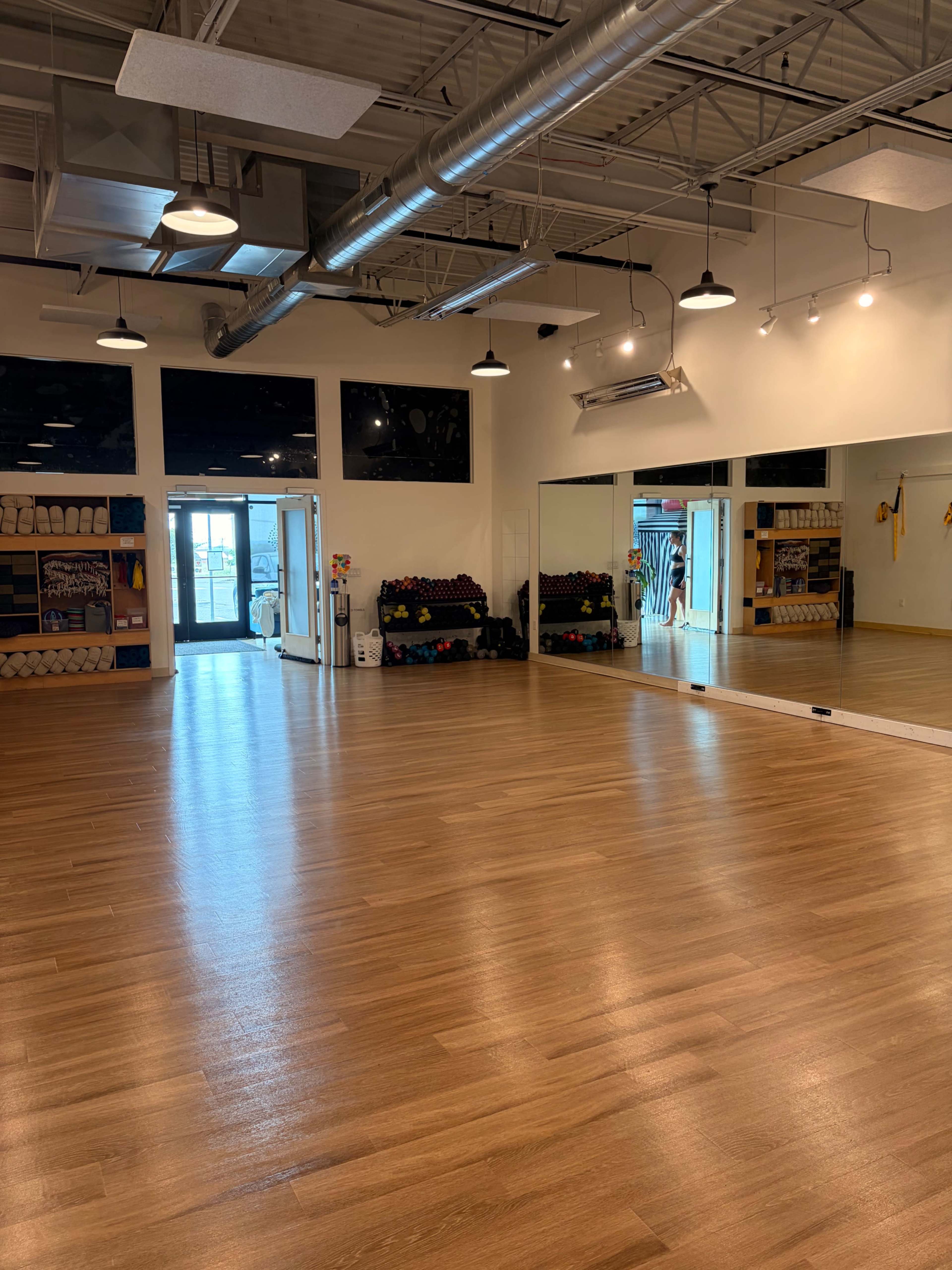 The image shows an empty exercise studio with wooden flooring, large mirrors, and workout equipment neatly organized along the walls.