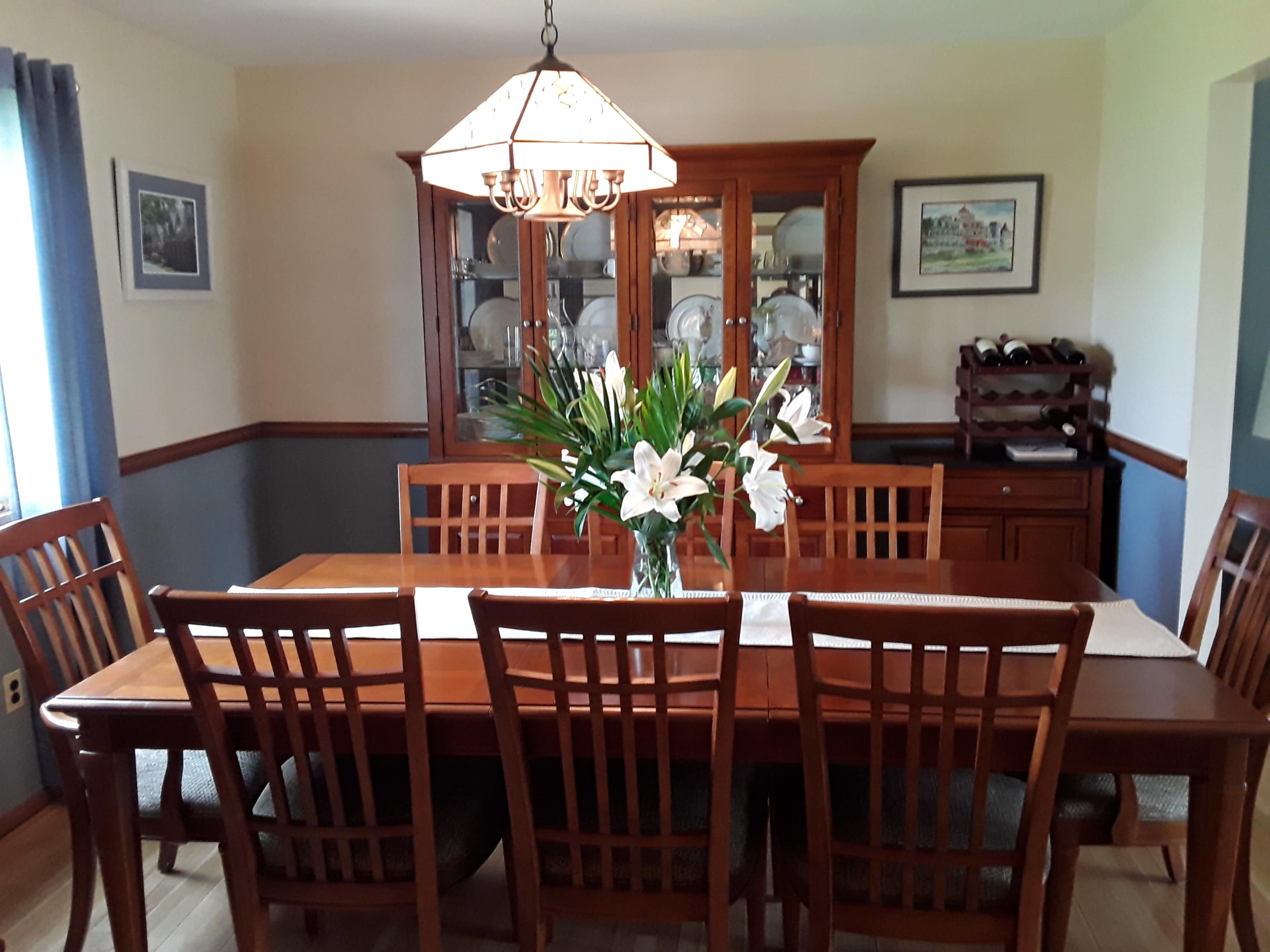 A wooden dining table is set at the center of a dining room, surrounded by chairs, with a vase of lilies on the table and a china cabinet in the background.