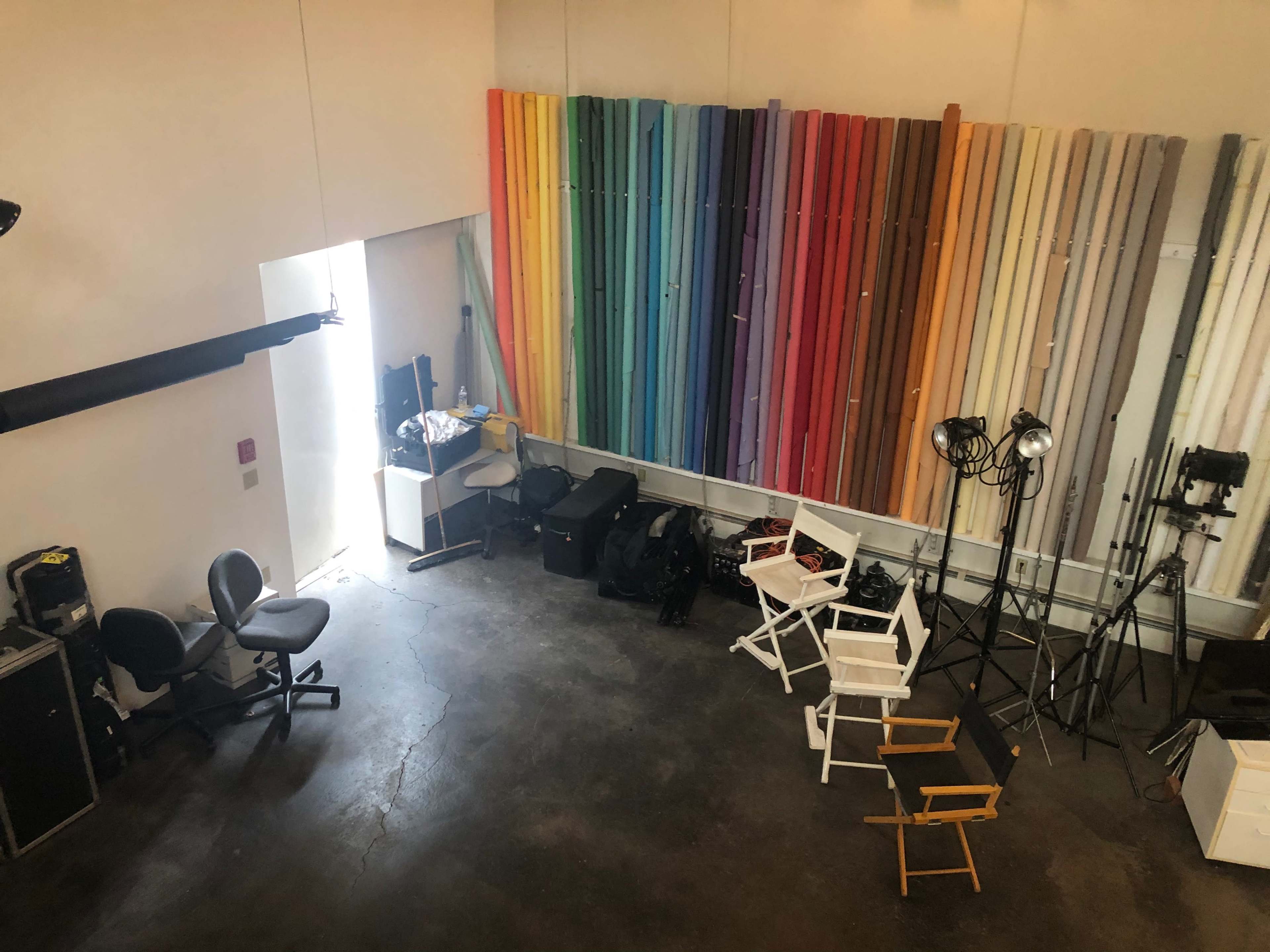 A photography studio with various colored backdrops arranged vertically on one wall, along with film equipment and director's chairs in the foreground.