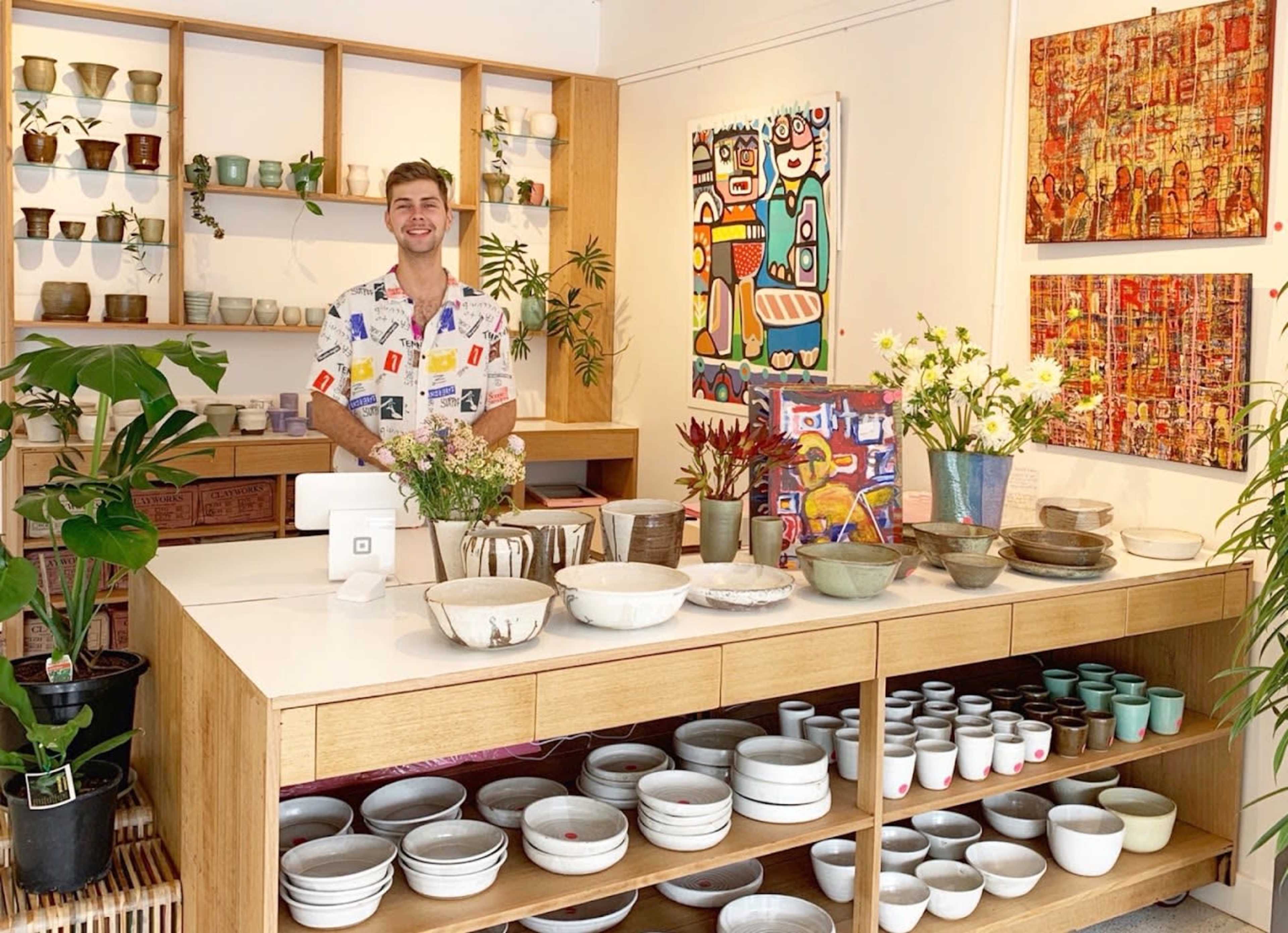 A bright gallery-style store features a smiling man standing behind a counter filled with various pottery and plants, while colorful artwork adorns the walls.