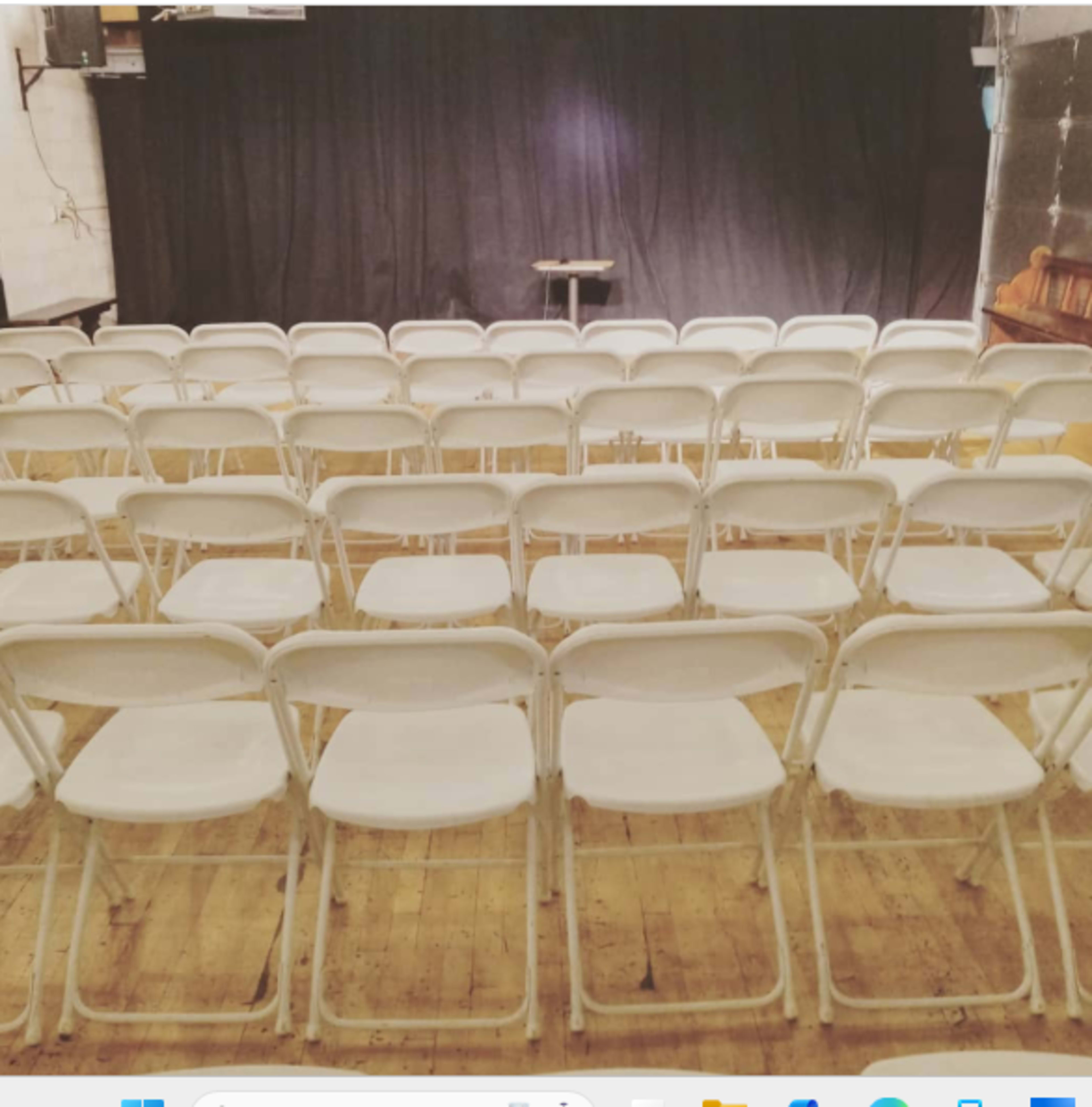The image shows a set of empty white chairs arranged in rows facing a stage with a black curtain in a performance venue.