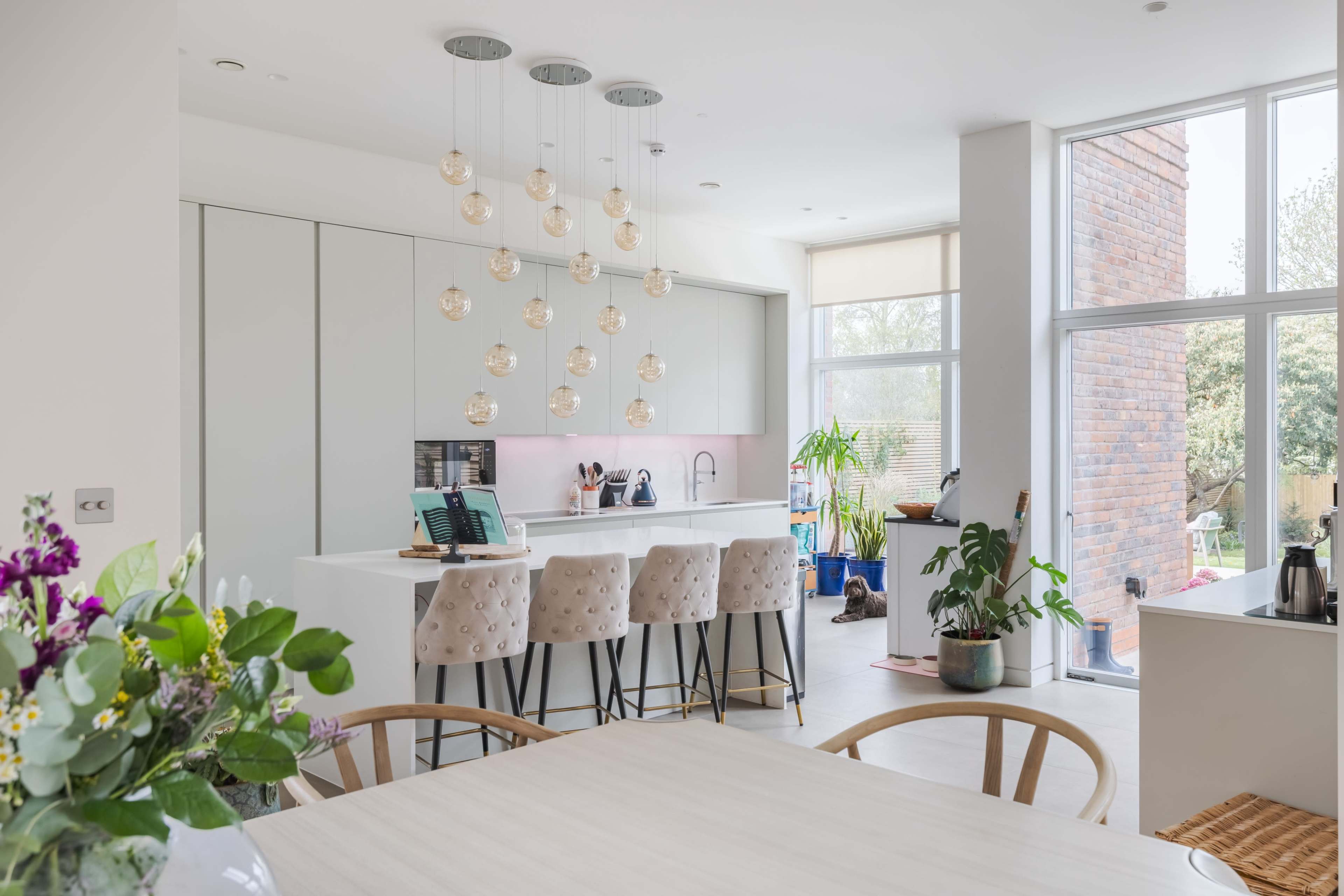 The image shows a modern kitchen with a large island, pendant lighting, and views of a garden through large windows.