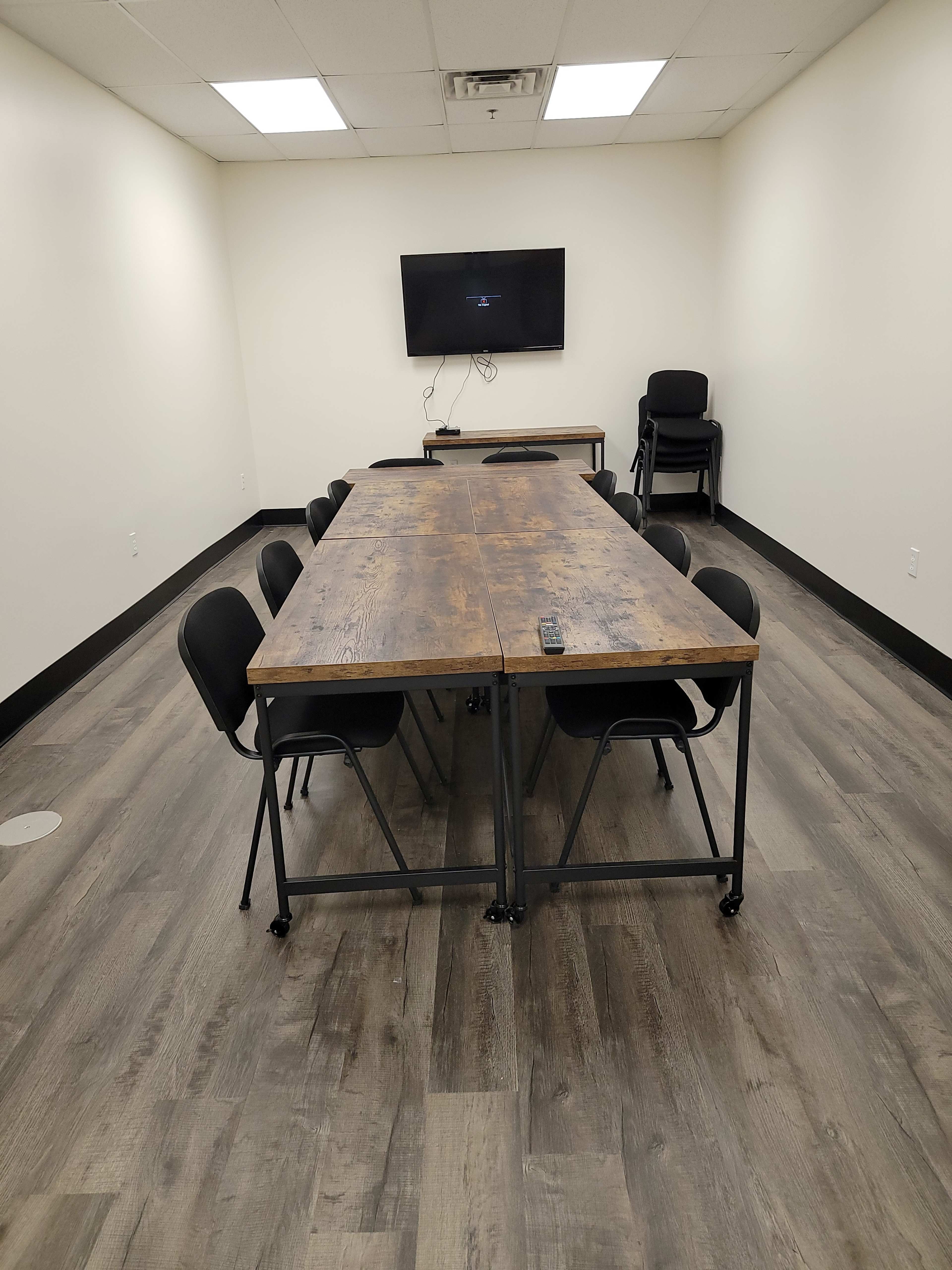 A long wooden table with chairs is set up in a minimally furnished meeting room featuring a television mounted on the wall.