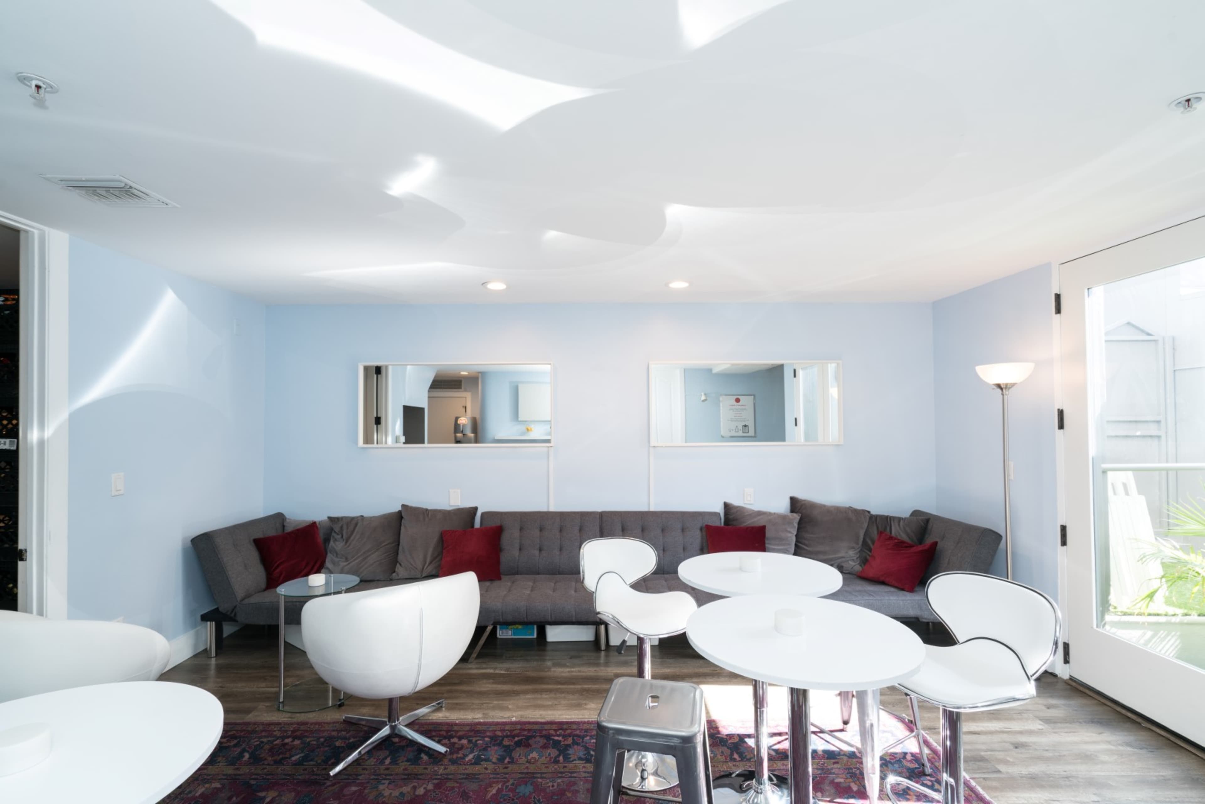 A modern lounge area with a gray sectional sofa, white chairs, round tables, and two mirrors on the wall, illuminated by natural light from large windows.