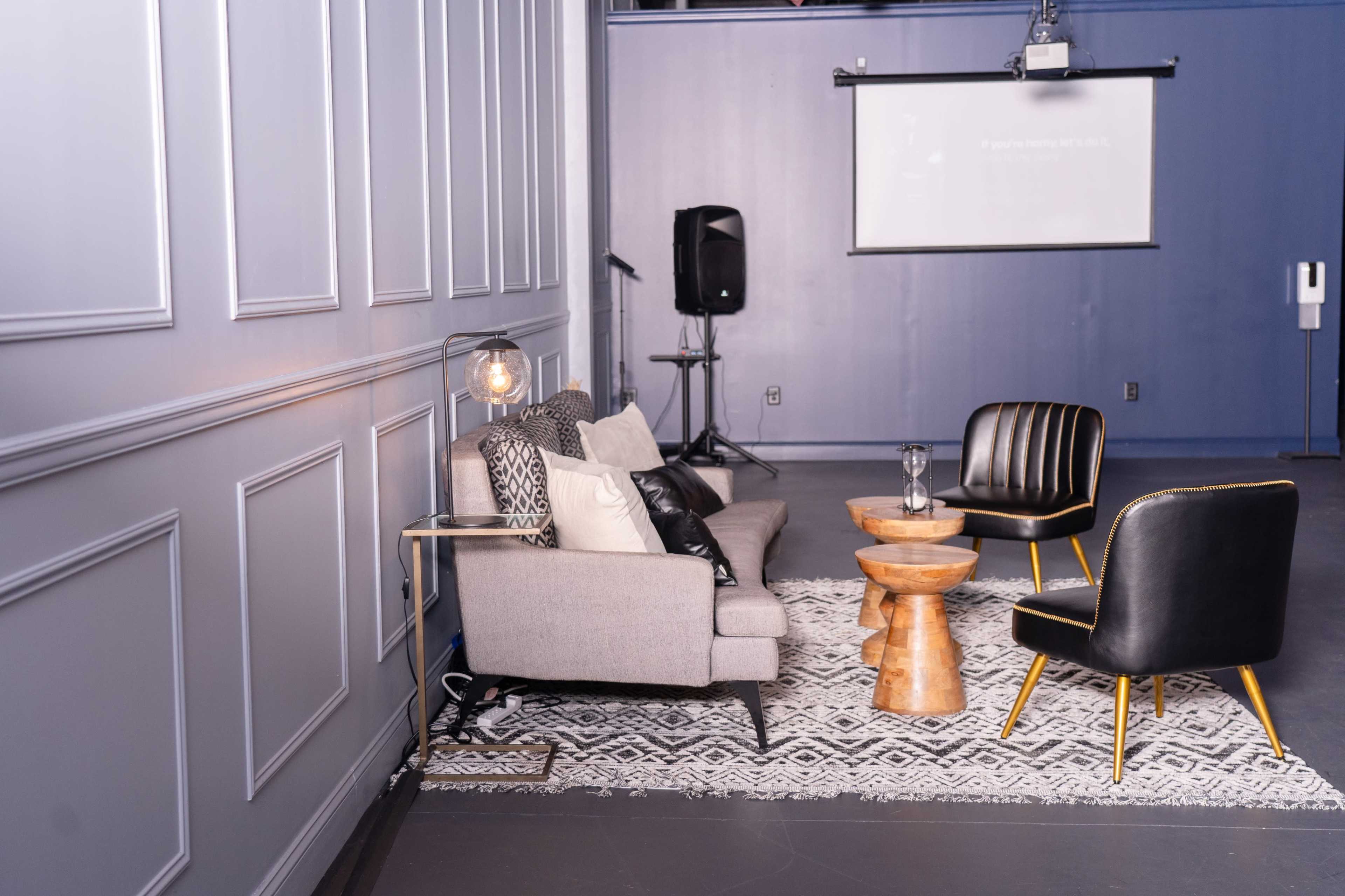 A stylish lounge area features a sofa, two black chairs, a wooden side table, and a projector screen in a modern space.
