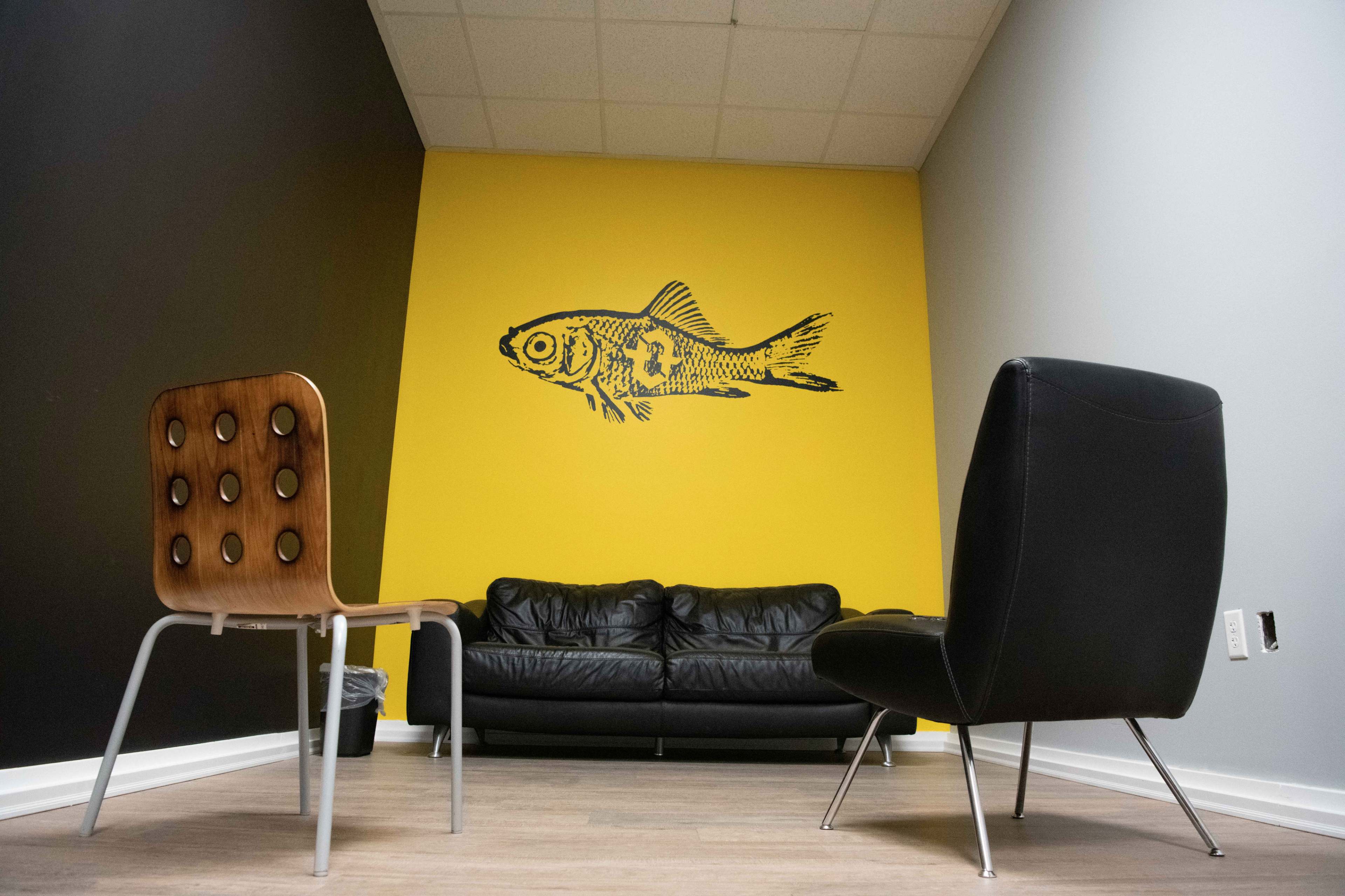 A waiting area features two chairs and a black sofa against a yellow wall decorated with a fish mural.