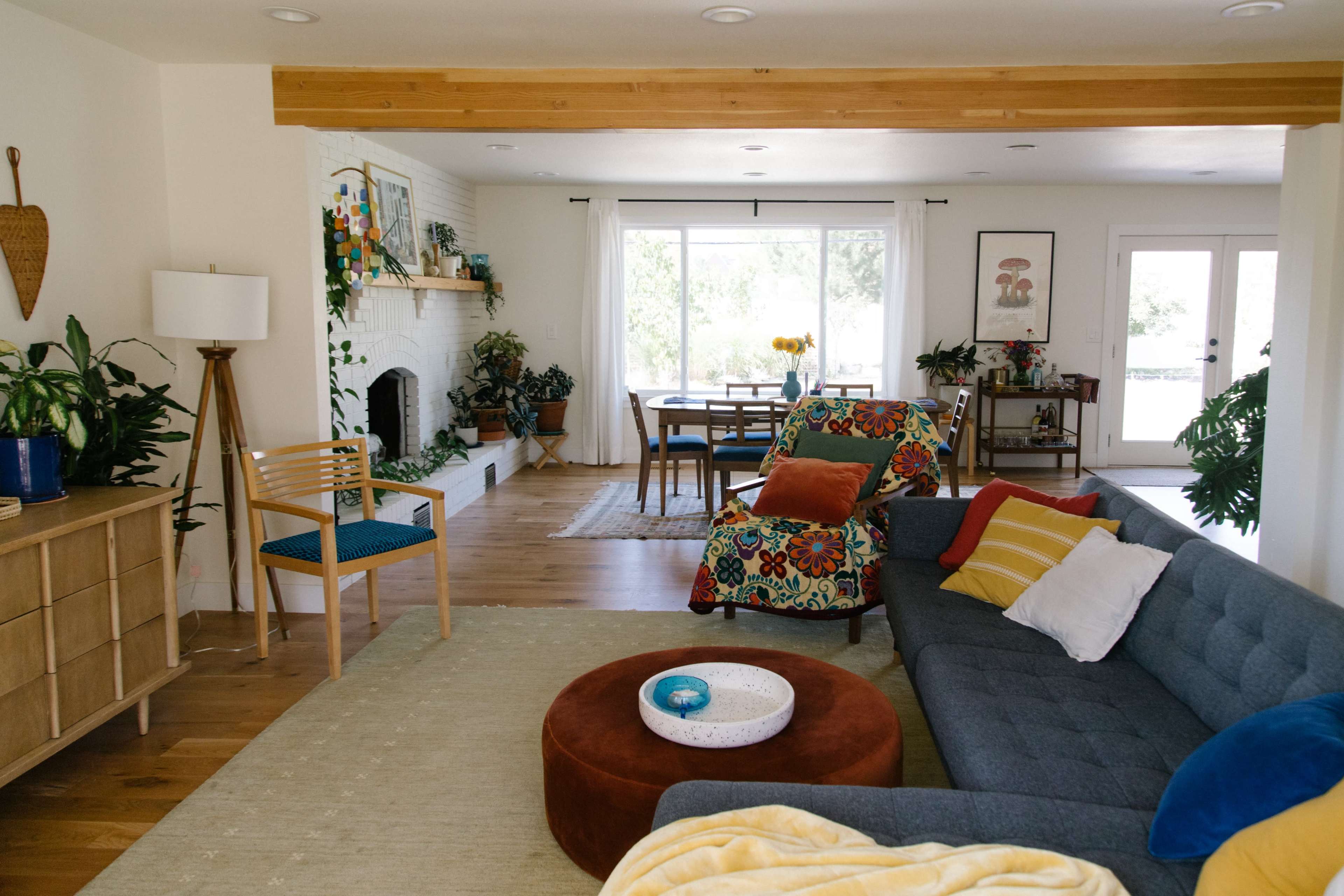 A bright living room with a sectional sofa, a wooden dining table, various plants, and a fireplace, all arranged in an open layout.