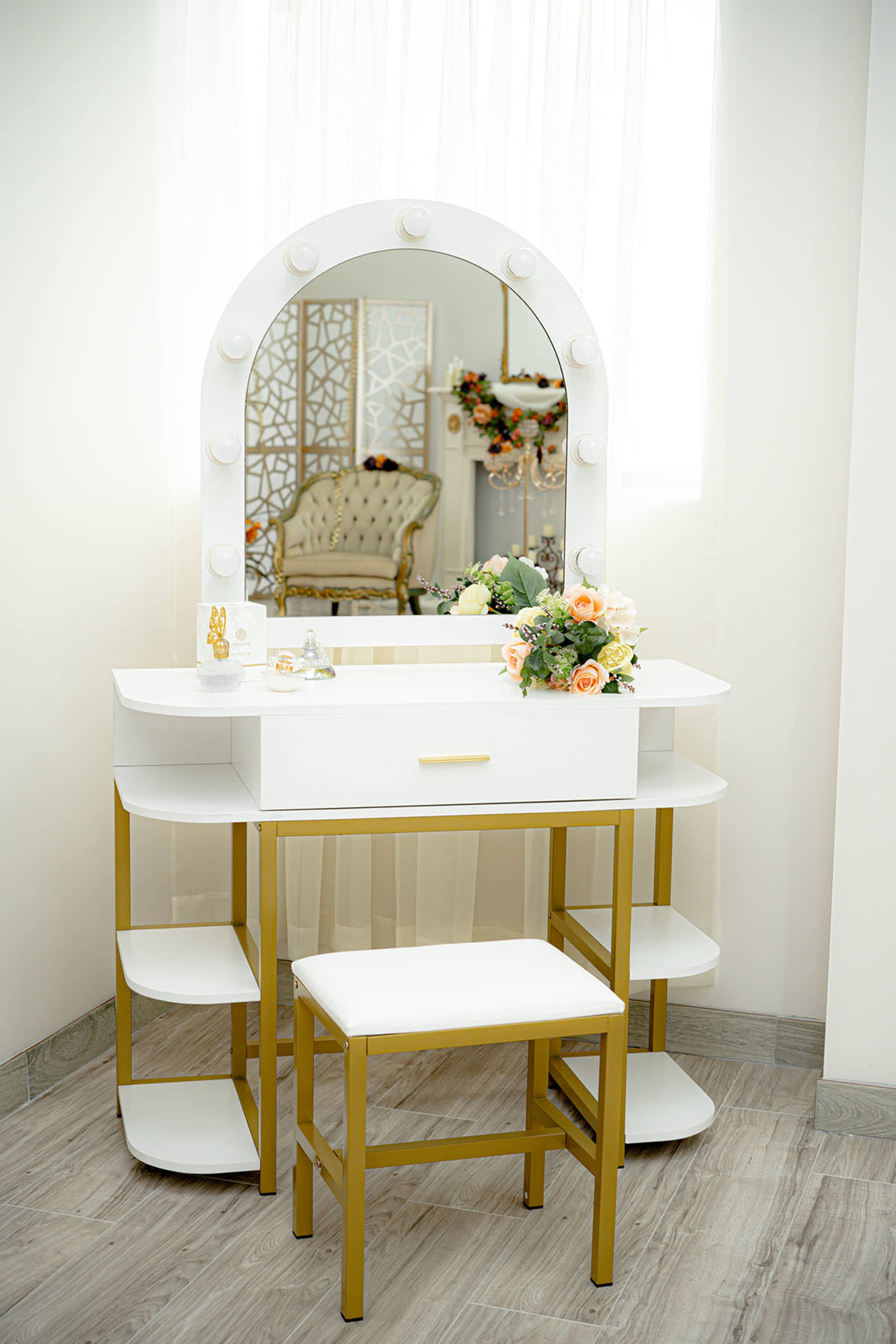 A modern vanity table with a large illuminated mirror, surrounded by decorative flowers and a small stool, is positioned in a bright room.