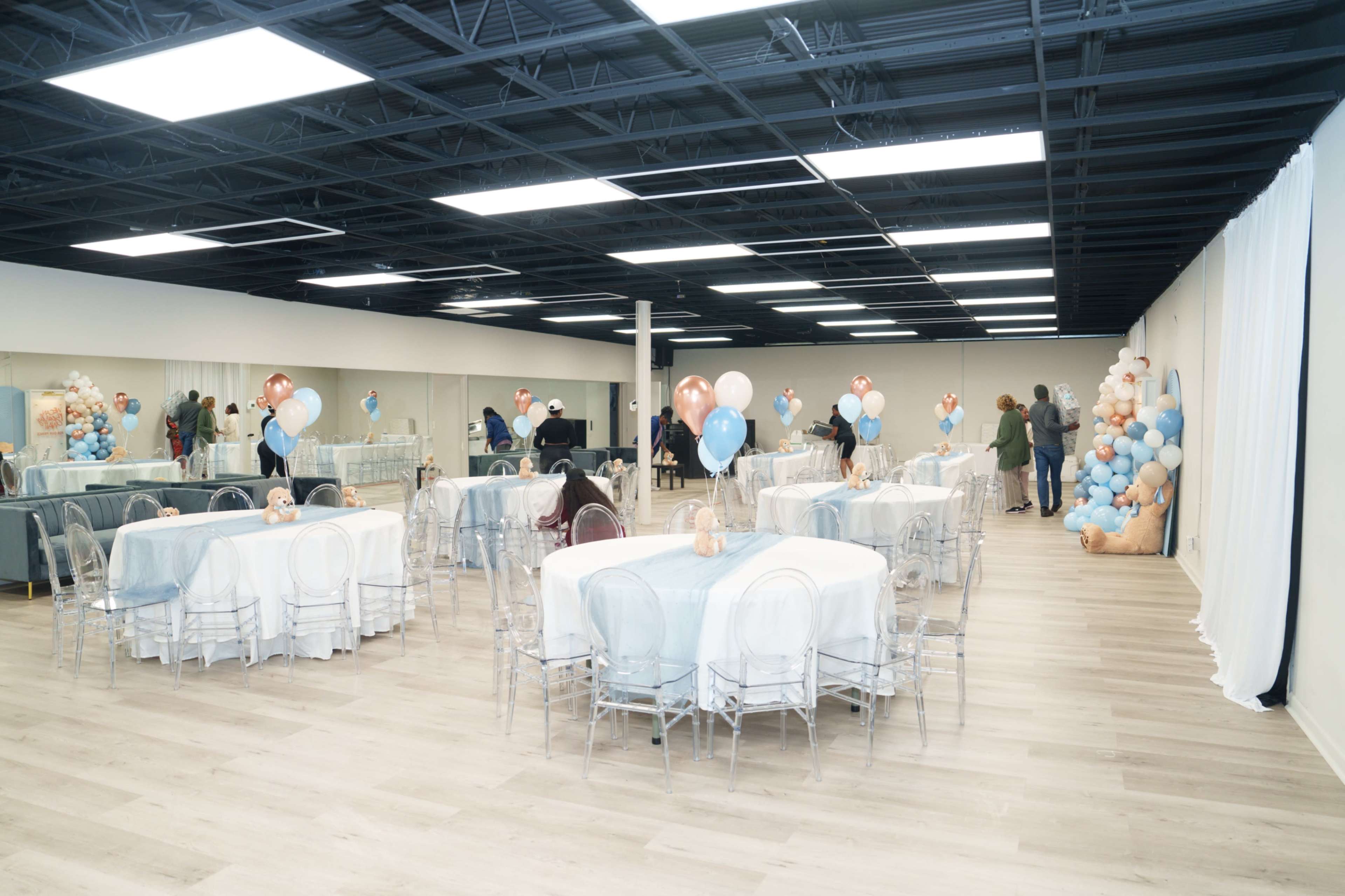 The image shows an event space with several round tables covered in white tablecloths, adorned with blue and peach balloons, and a large balloon arrangement in one corner.