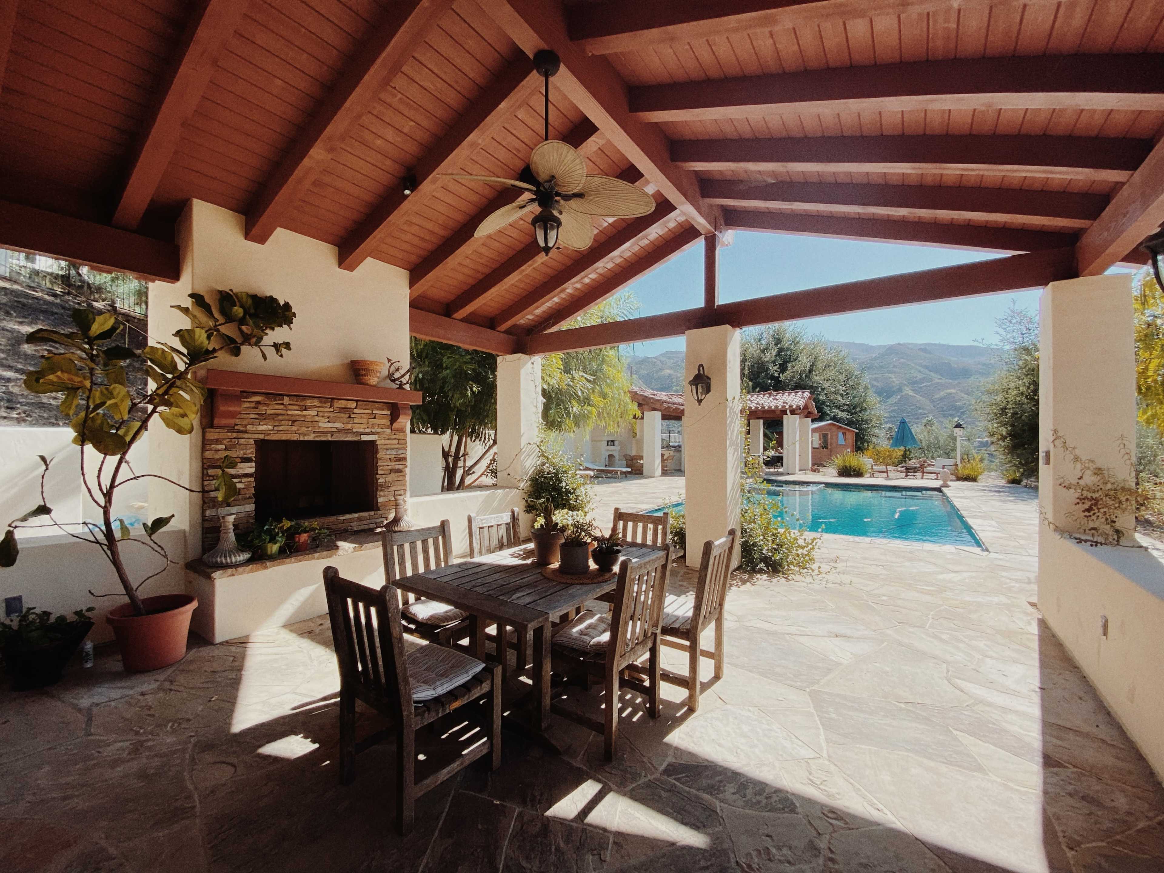 A covered patio features a wooden dining table surrounded by chairs, with a stone fireplace and a view of a swimming pool and mountains in the background.
