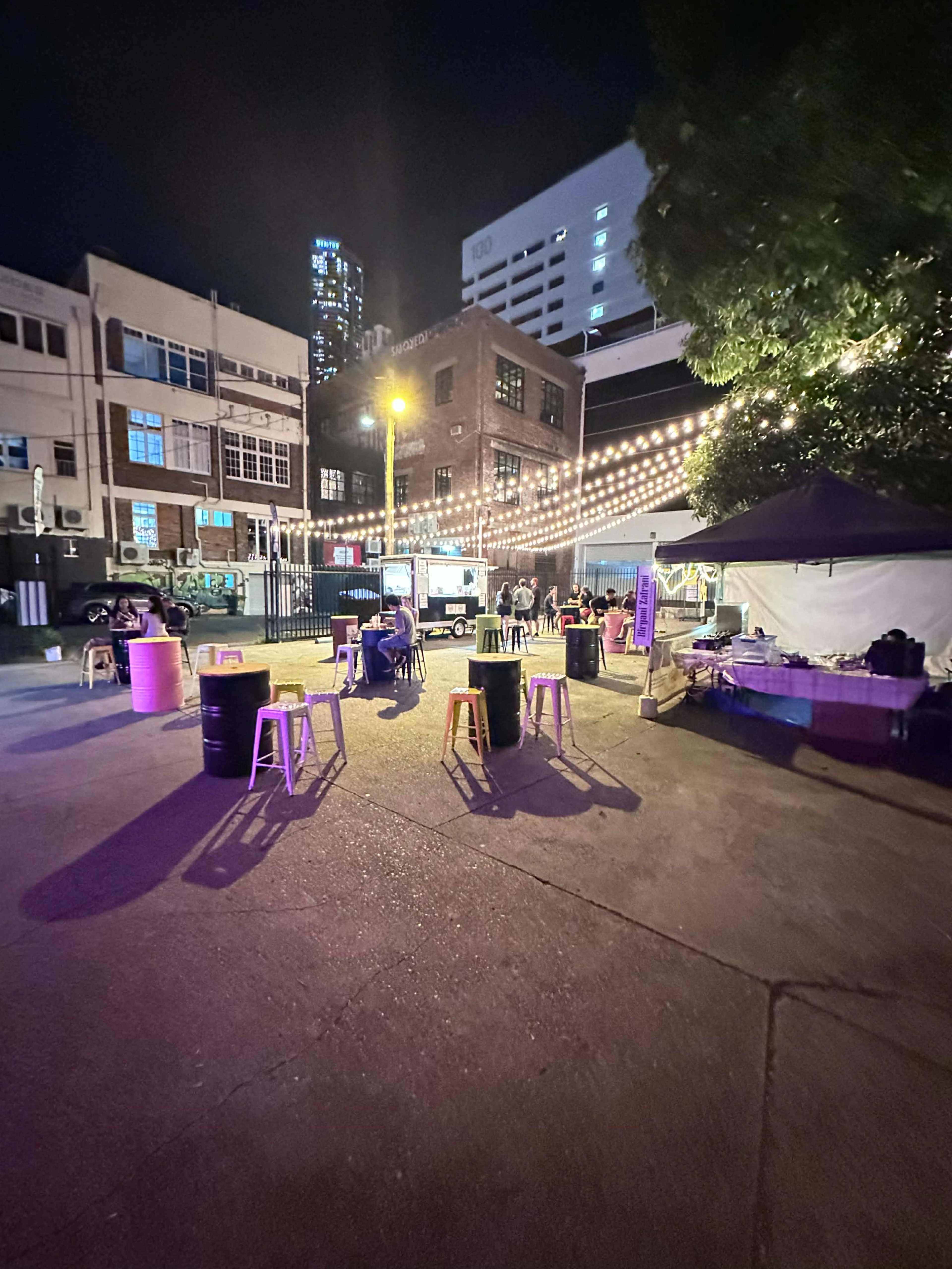 The scene shows a night-time outdoor gathering with lit string lights, tables, and people sitting around in an urban setting.