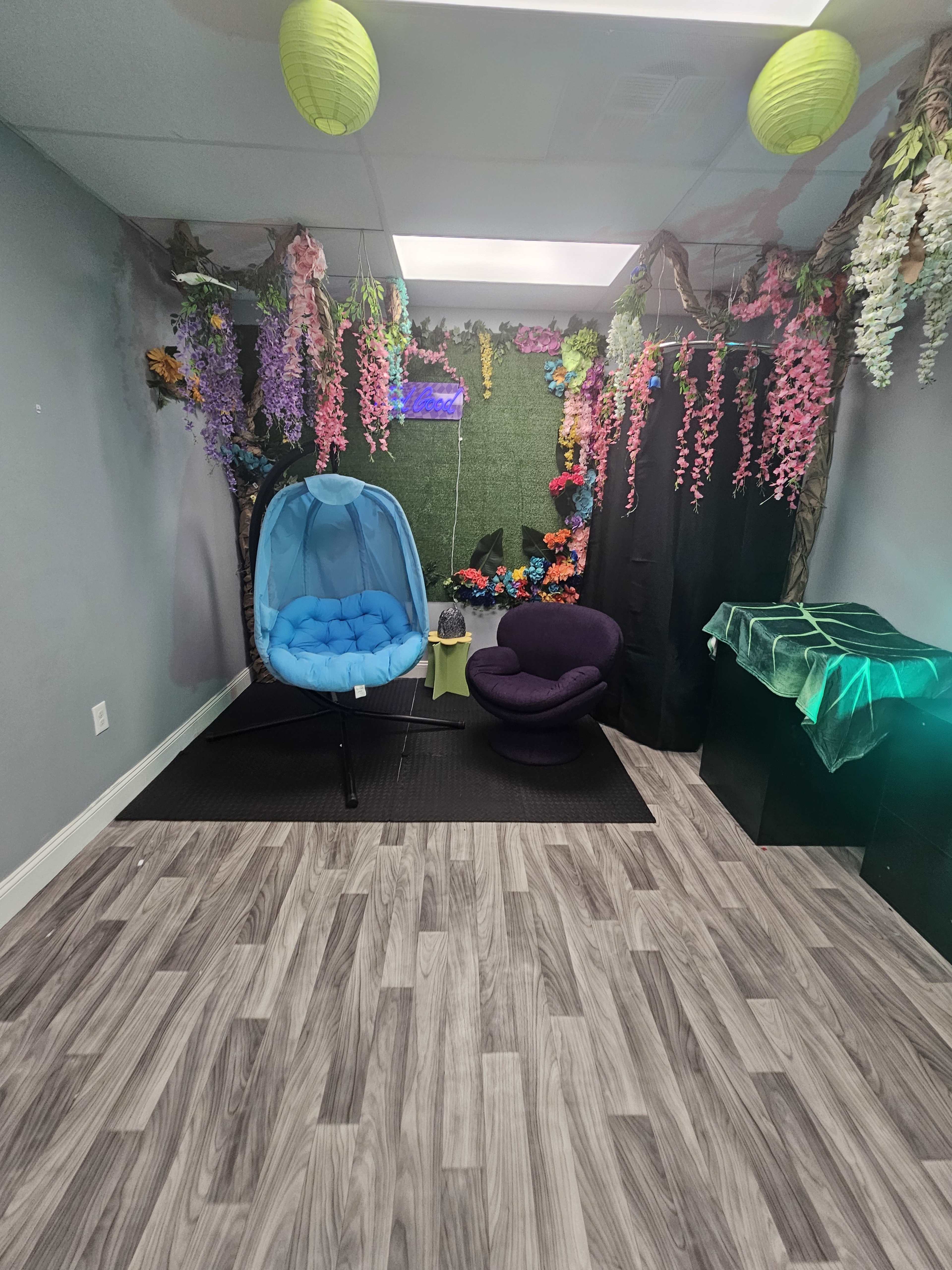 A room decorated with artificial flowers, green foliage, and colorful hanging lanterns, featuring a blue hanging chair and a purple chair on a wood-like floor.