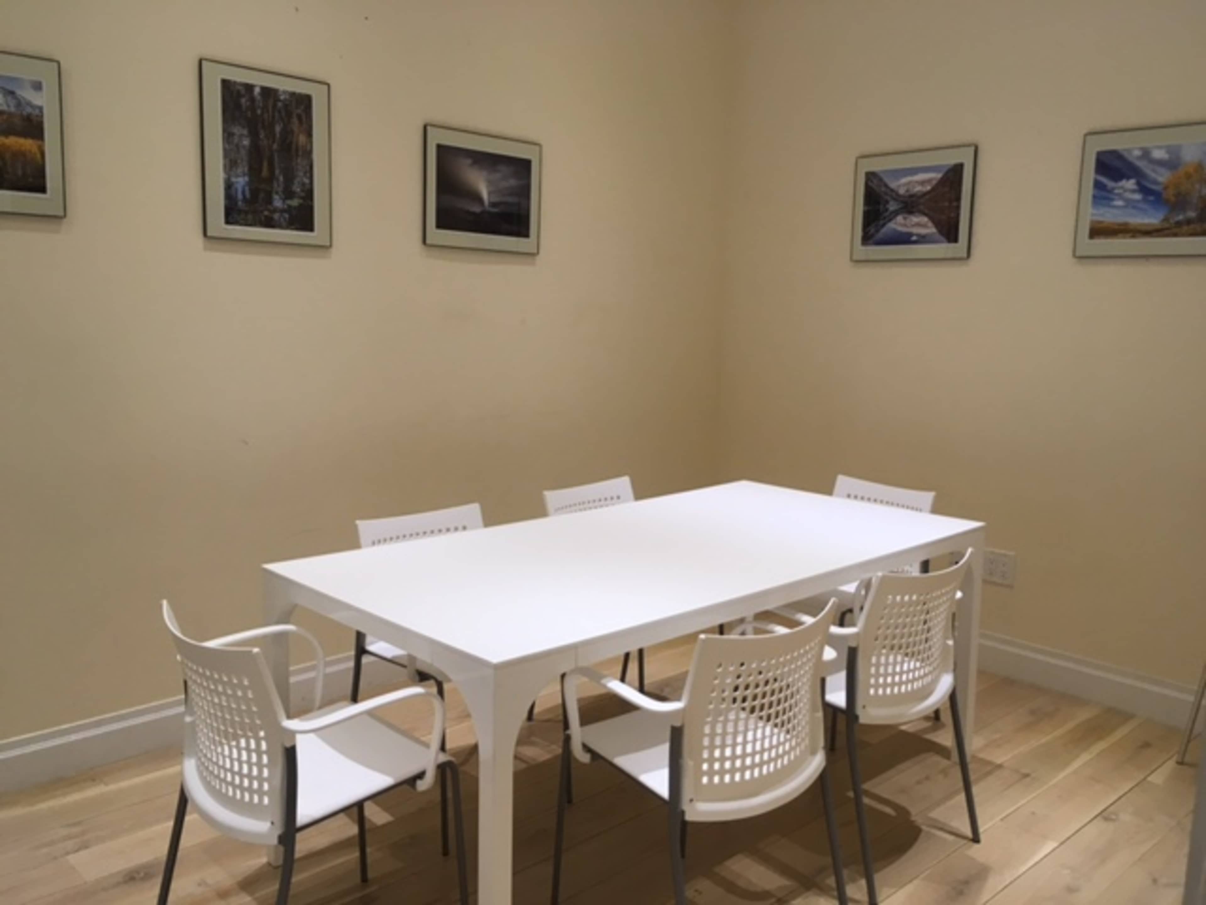 A rectangular white table is surrounded by six white chairs in a room with light-colored walls and framed landscape photographs on the walls.