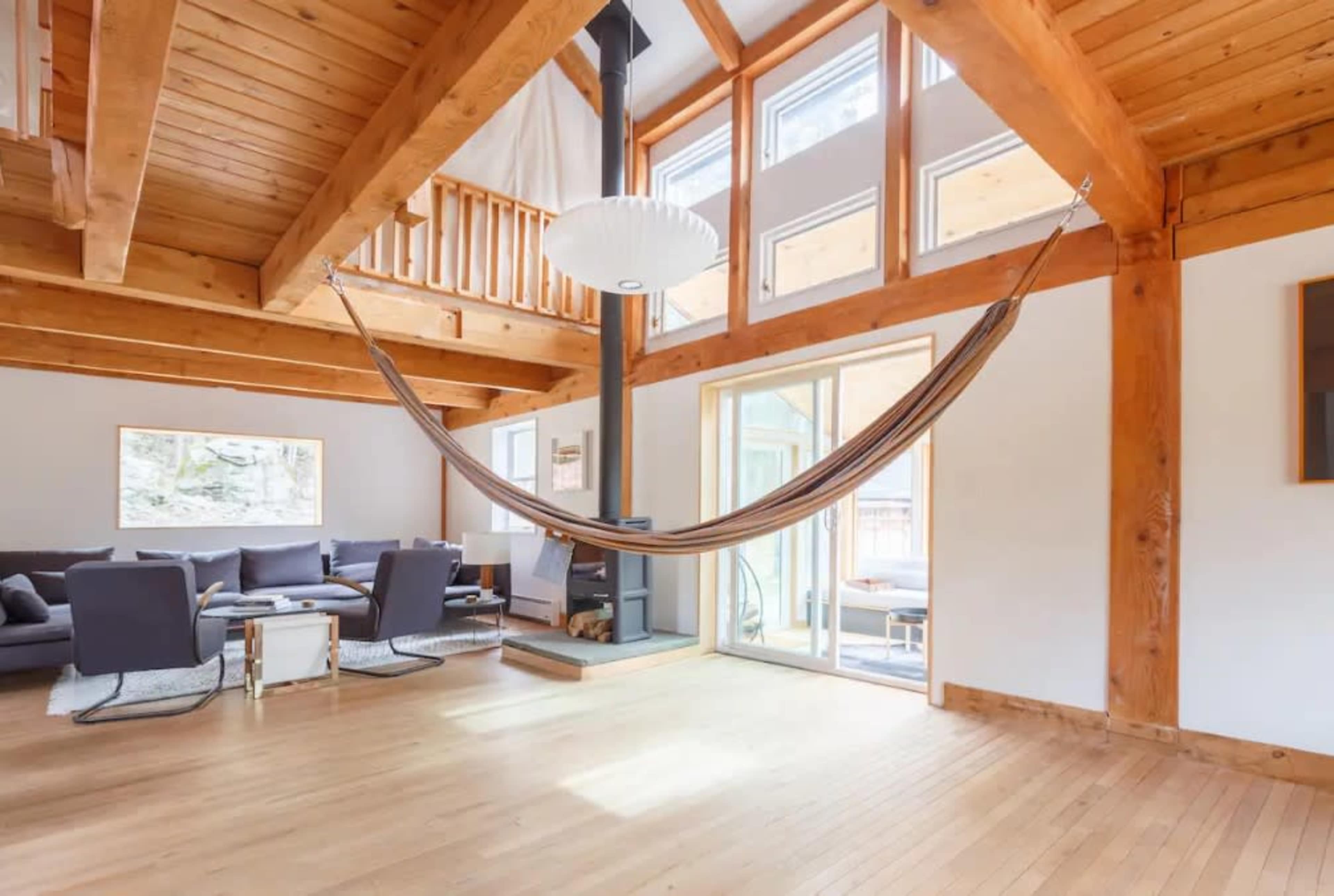 The image shows a spacious living room with wooden beams, a hammock suspended in the center, and large windows allowing natural light to enter.