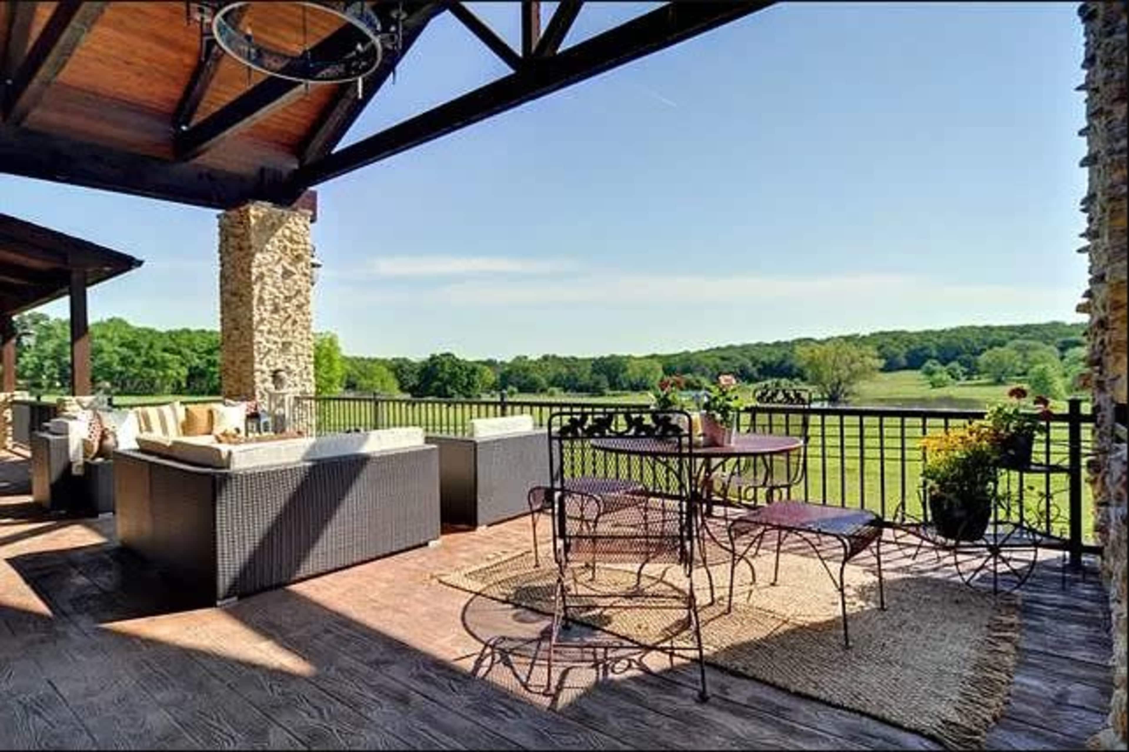 A covered patio features a seating area with wicker furniture and a dining table, overlooking a lush green landscape.