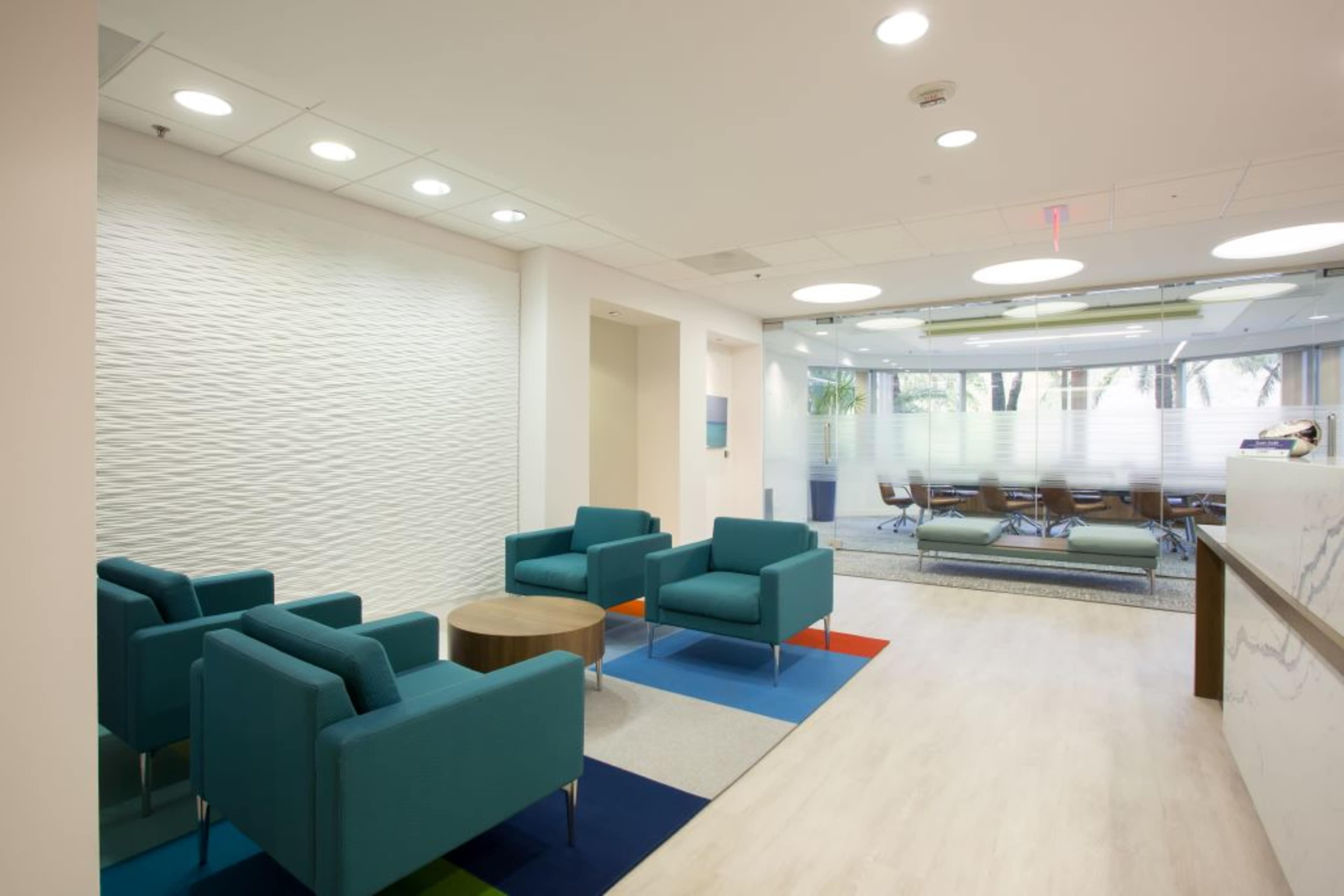 The image shows a modern office lobby with teal chairs arranged around a small wooden table, adjacent to a conference room with large windows.