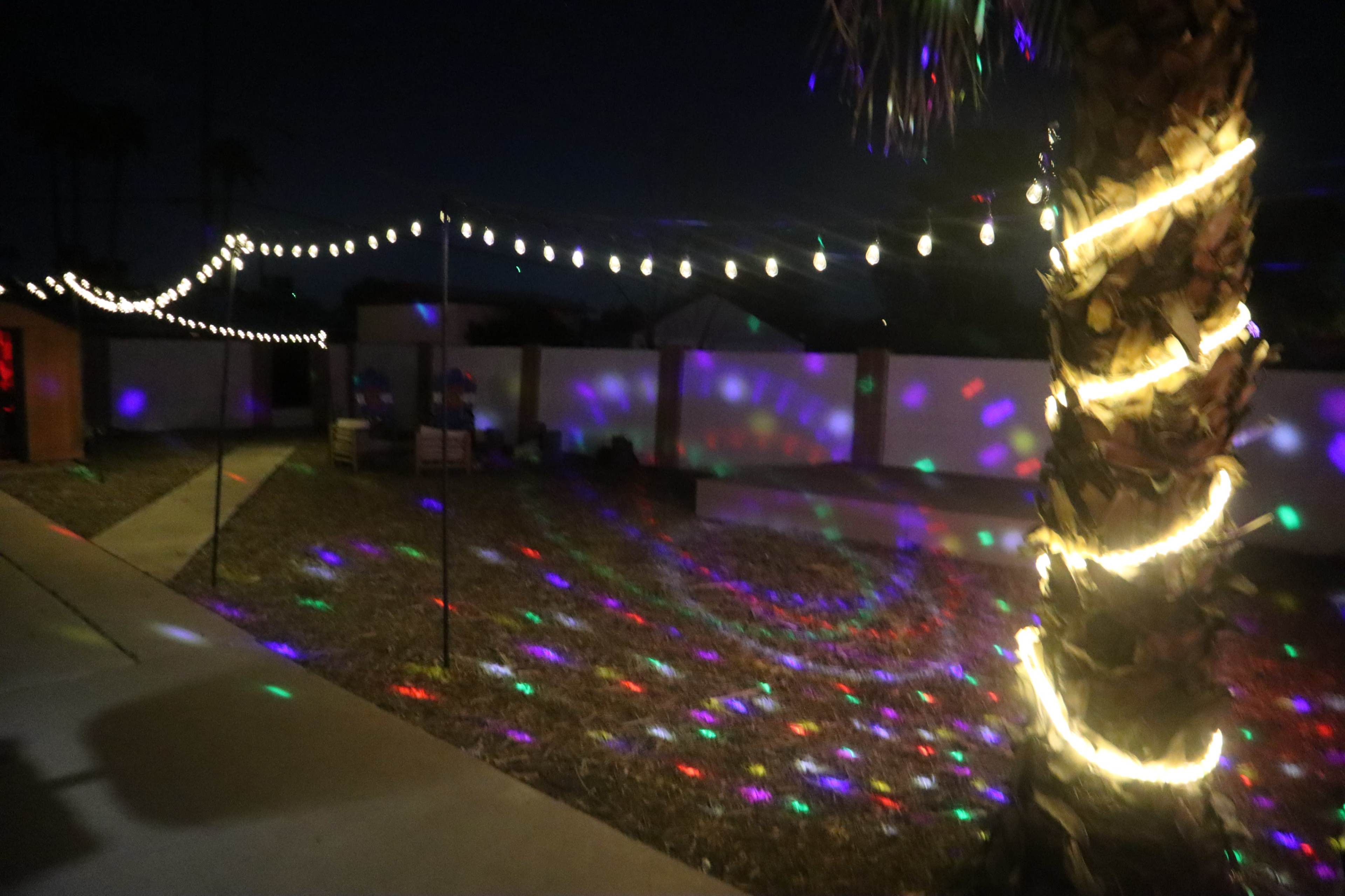 The image shows a backyard at night, illuminated by string lights and colorful projections on the ground and walls.