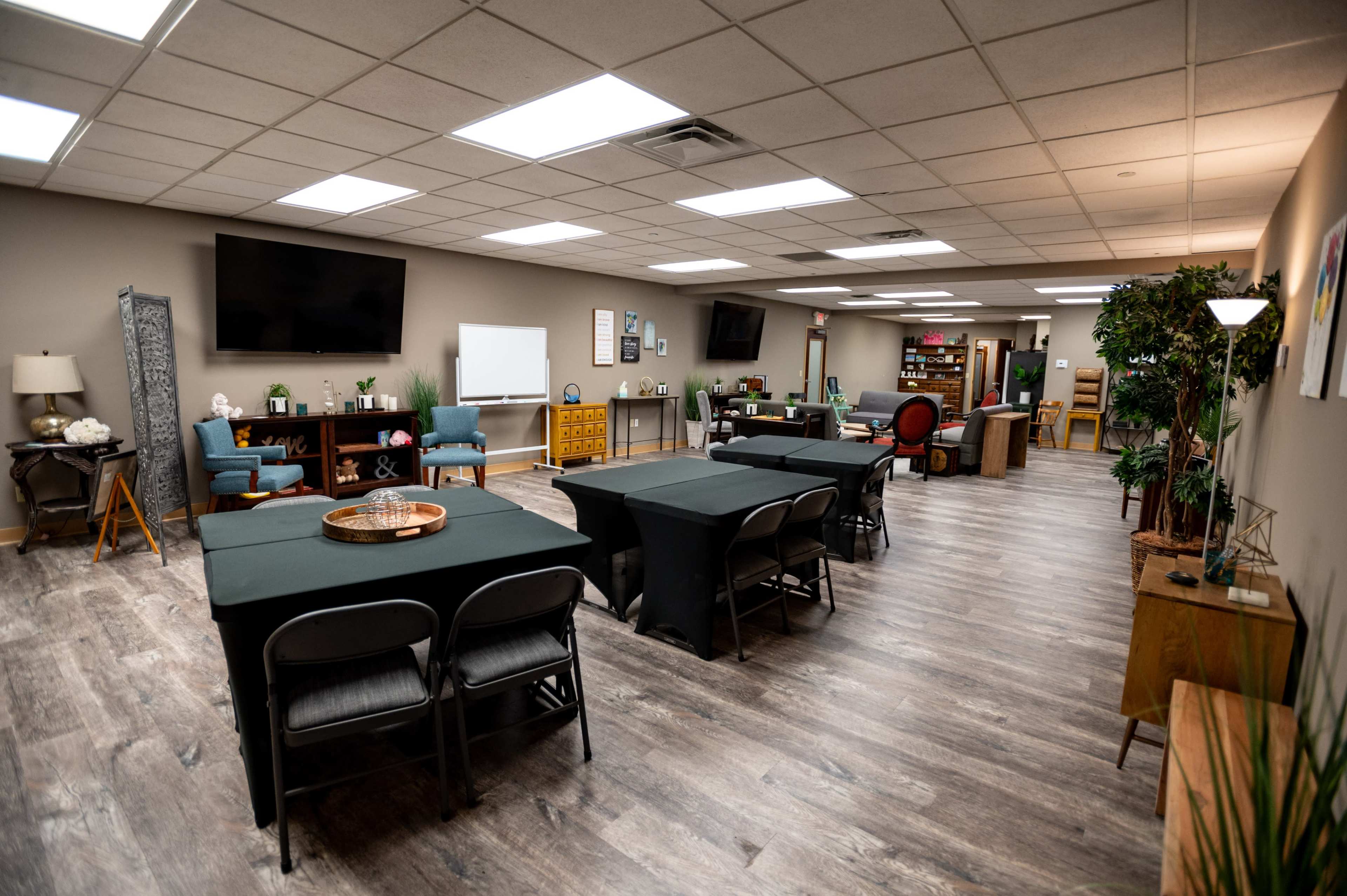 A well-lit meeting room with multiple black-covered tables, two large televisions on the walls, and various seating and decor items.