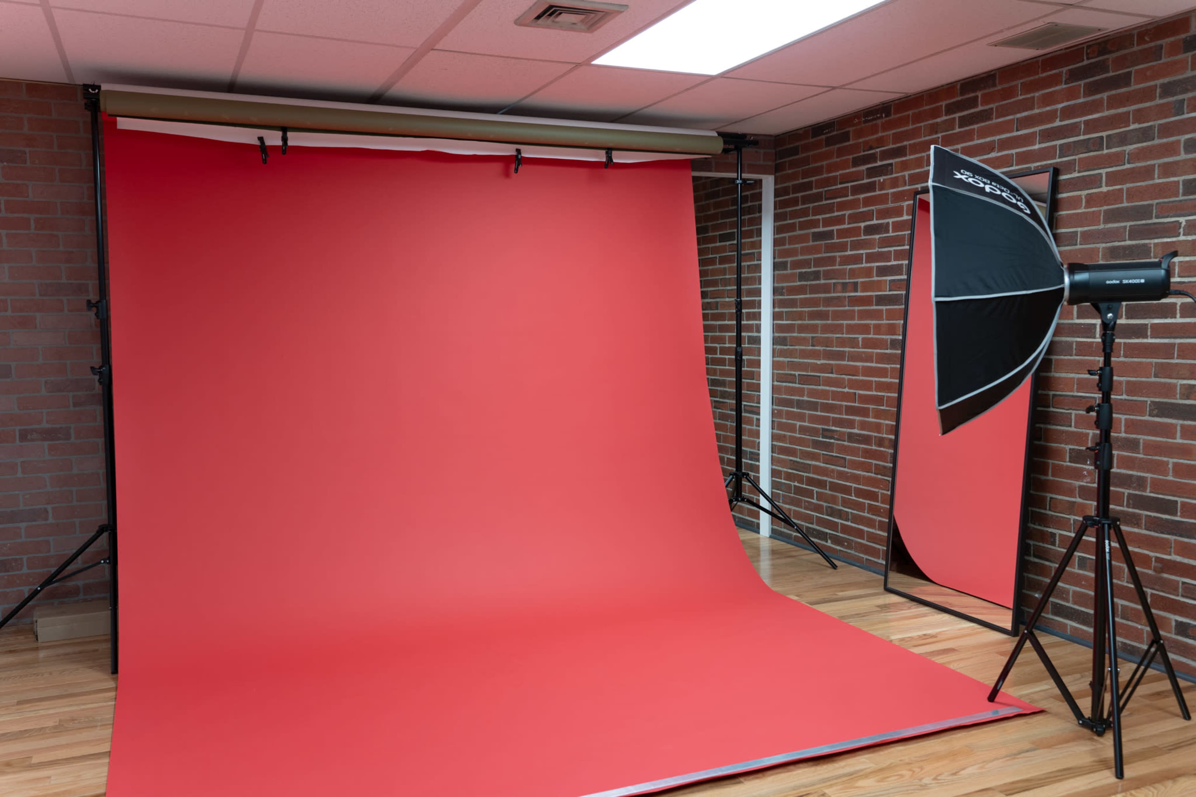 The image shows a photography studio with a red backdrop, wooden flooring, and a softbox light setup on the side.