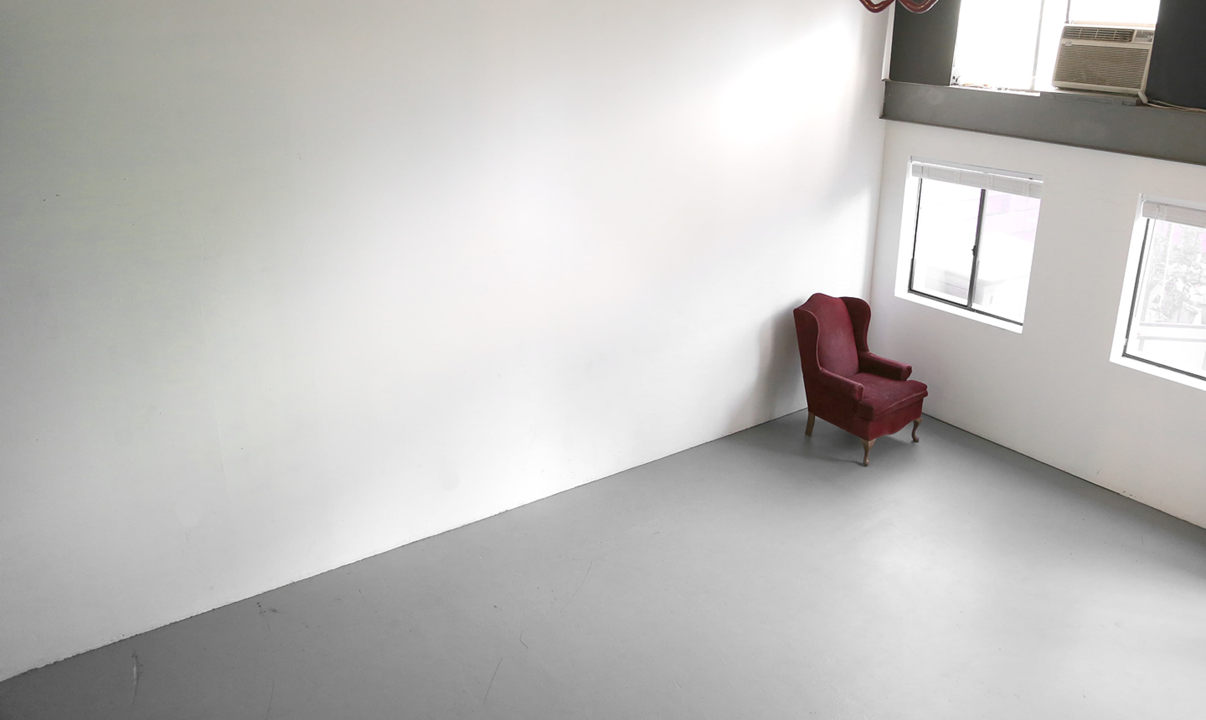 An empty room features a single red armchair positioned against a white wall, with two windows allowing natural light.